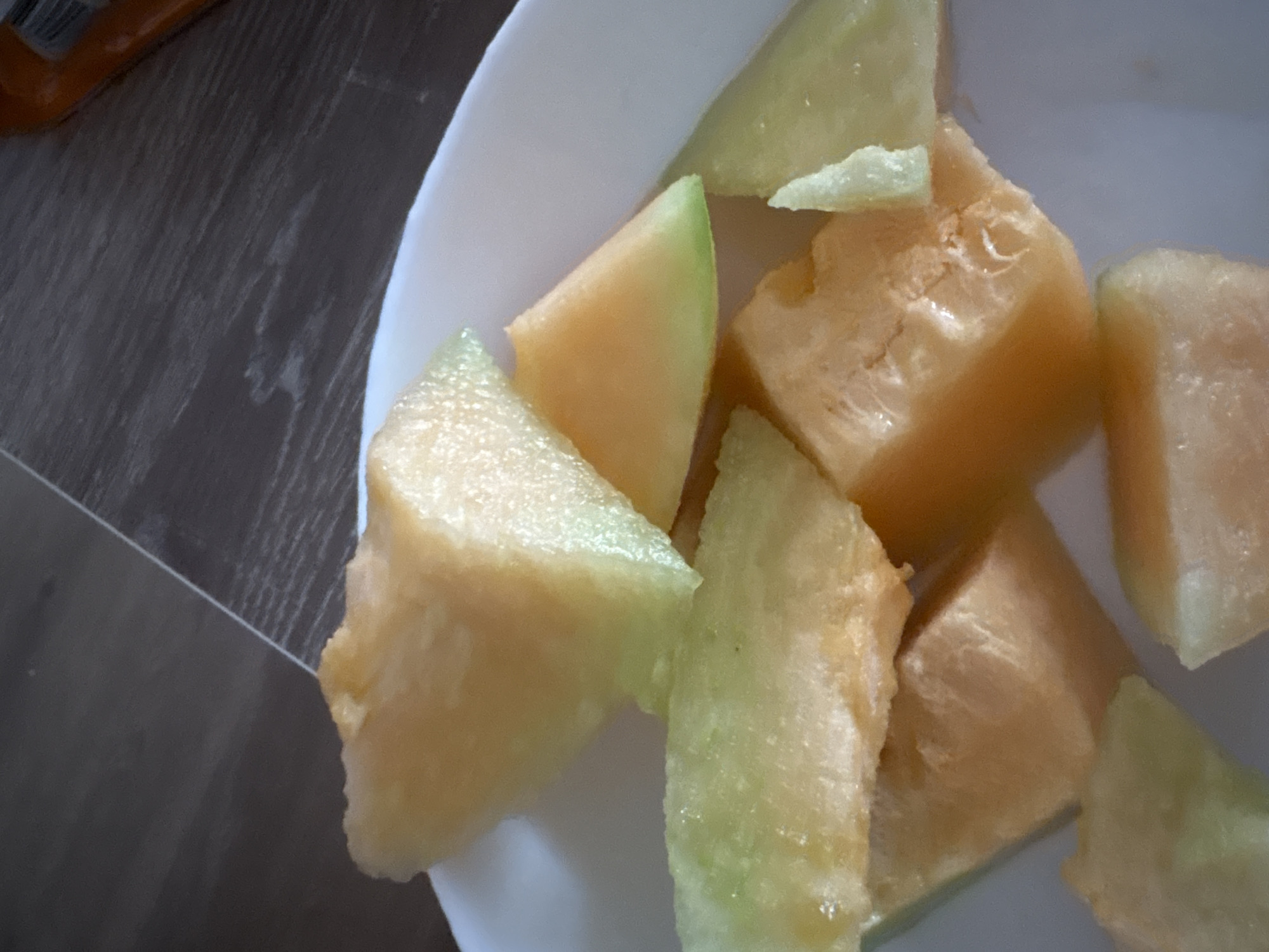 A white bowl filled with chopped cantaloupe sits on a wooden floor. The melon pieces are a mixture of orange and pale green. The lighting is natural, casting soft shadows. The overall impression is a simple, domestic scene.