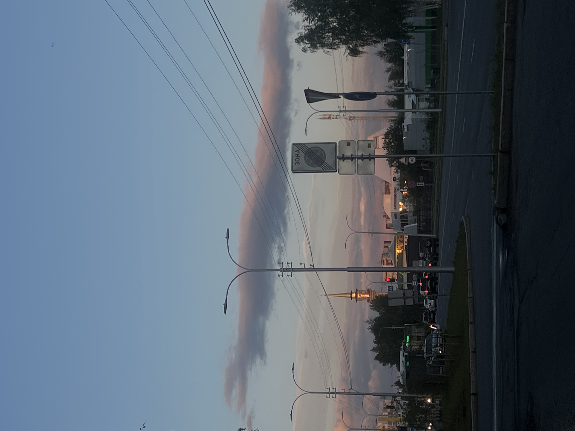 The image depicts a twilight scene on a street in Surgut, Russia. A tall, golden-domed building, likely a mosque or church with a minaret, is visible in the background, illuminated by the setting sun. Several streetlights line the road, and overhead power lines crisscross the sky. Traffic lights show red, indicating vehicles are stopped. Various cars and a large truck are present on the road. Buildings and trees are visible along the sides of the street. The sky is a soft gradient of blue and pink from the sunset. A traffic sign with the Cyrillic word "ЗОНА" and a circular emblem indicating a restricted area is prominent in the middle ground. Another sign below it is also visible. The overall impression is that of a busy urban intersection at dusk.