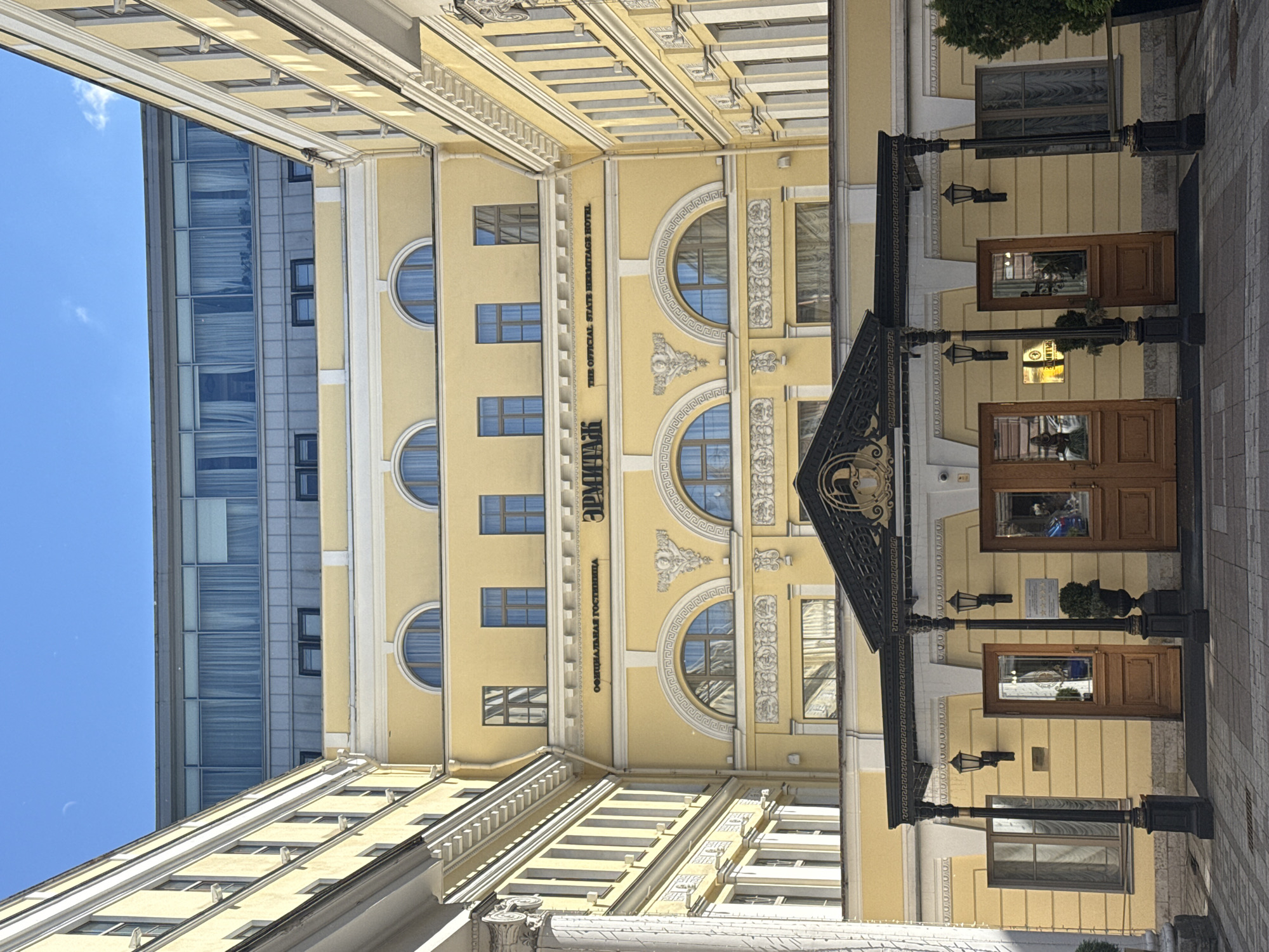 The image shows the facade of The Official State Hermitage Hotel in Saint Petersburg, Russia. The hotel is a yellow building with classical architectural elements, featuring arched windows, ornate decorations, and a prominent entrance with a black awning supported by decorative pillars. The building's upper floors are topped by a more modern structure with large windows. The sky is clear and blue, suggesting daytime. The scene is devoid of people, and the focus is on the architectural details of the hotel.