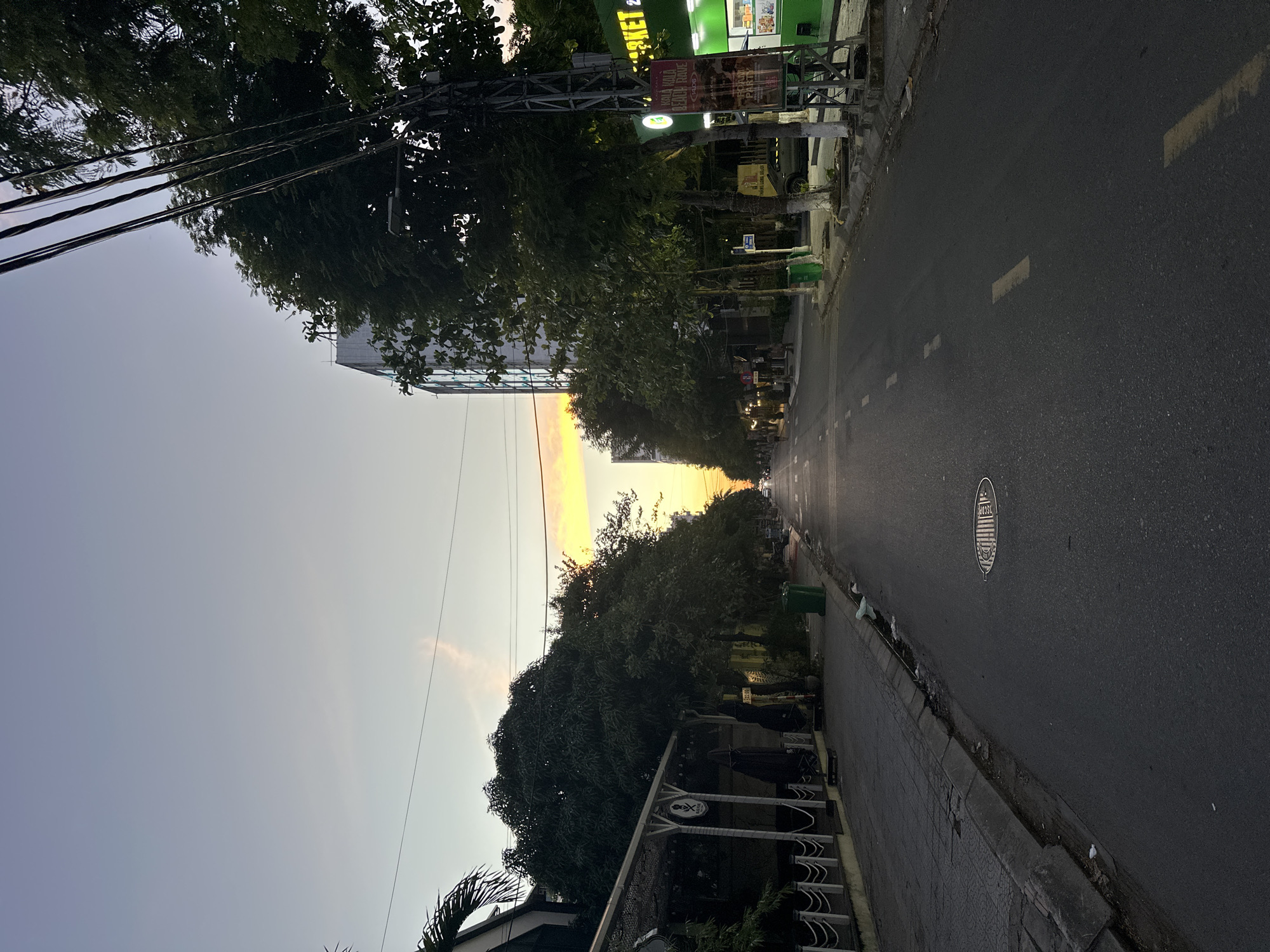 This image depicts a street scene in Da Nang, Vietnam, during what appears to be dusk or early morning. The perspective is from the middle of the road, looking down the street. The sky is a soft gradient of pale blue to a warm orange glow on the horizon, indicating either sunrise or sunset.
On the left side of the street, there is a sidewalk with a tiled surface, bordered by a low white fence. Adjacent to the sidewalk is a building with an outdoor seating area, covered by a translucent canopy. Some tables and chairs are visible, along with signage that includes a skull and crossbones logo and text that may be in Vietnamese. Trees with dense foliage line the edge of the sidewalk. A green trash can is also present.
The road itself is asphalt, with faded yellow lane markings. A manhole cover with a circular pattern is visible in the foreground. On the right side of the road, there is another sidewalk with trees, a utility pole with numerous wires, and a brightly lit green building with signage that includes "MARKET" in large letters. Another sign on the utility pole advertises "KOLA KOLA BEACH RAVE." A few people are walking on the sidewalk in the distance. The overall impression is of a residential or commercial street with urban infrastructure, bathed in the soft light of twilight.