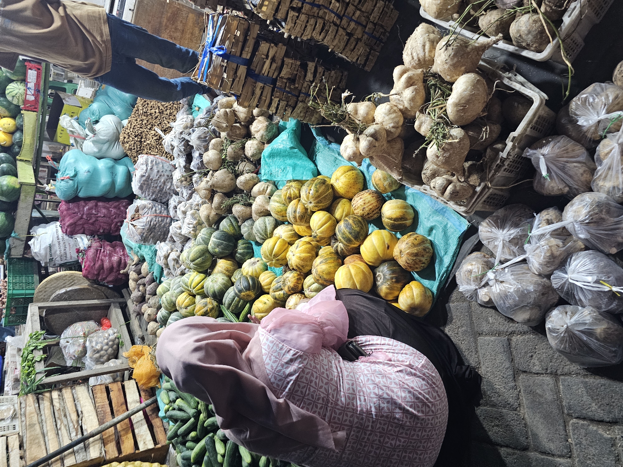 The image shows an outdoor market stall in Sidoharjo, Indonesia. In the foreground, a person wearing a pink hijab and a patterned pink and white shirt is kneeling, facing away from the camera. They are next to a large pile of small, round, yellow and green squashes. To the left of the squashes, there is a pile of cucumbers. In the background, there are sacks of produce, wooden crates, and other market goods. The ground is paved with dark, irregular stones. The lighting suggests it is either early morning or evening.
