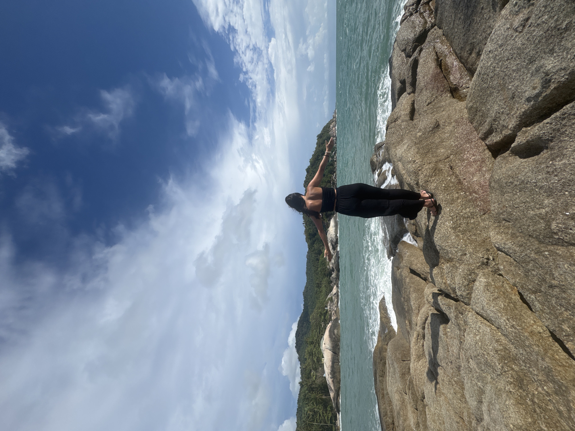 A woman stands on a rocky outcrop overlooking the ocean in Baan Thong Nang, Thailand. She is wearing a black top and pants and her arms are outstretched. The sky is blue with white clouds. The water is a light turquoise color. Trees cover a large rock formation along the shoreline.
