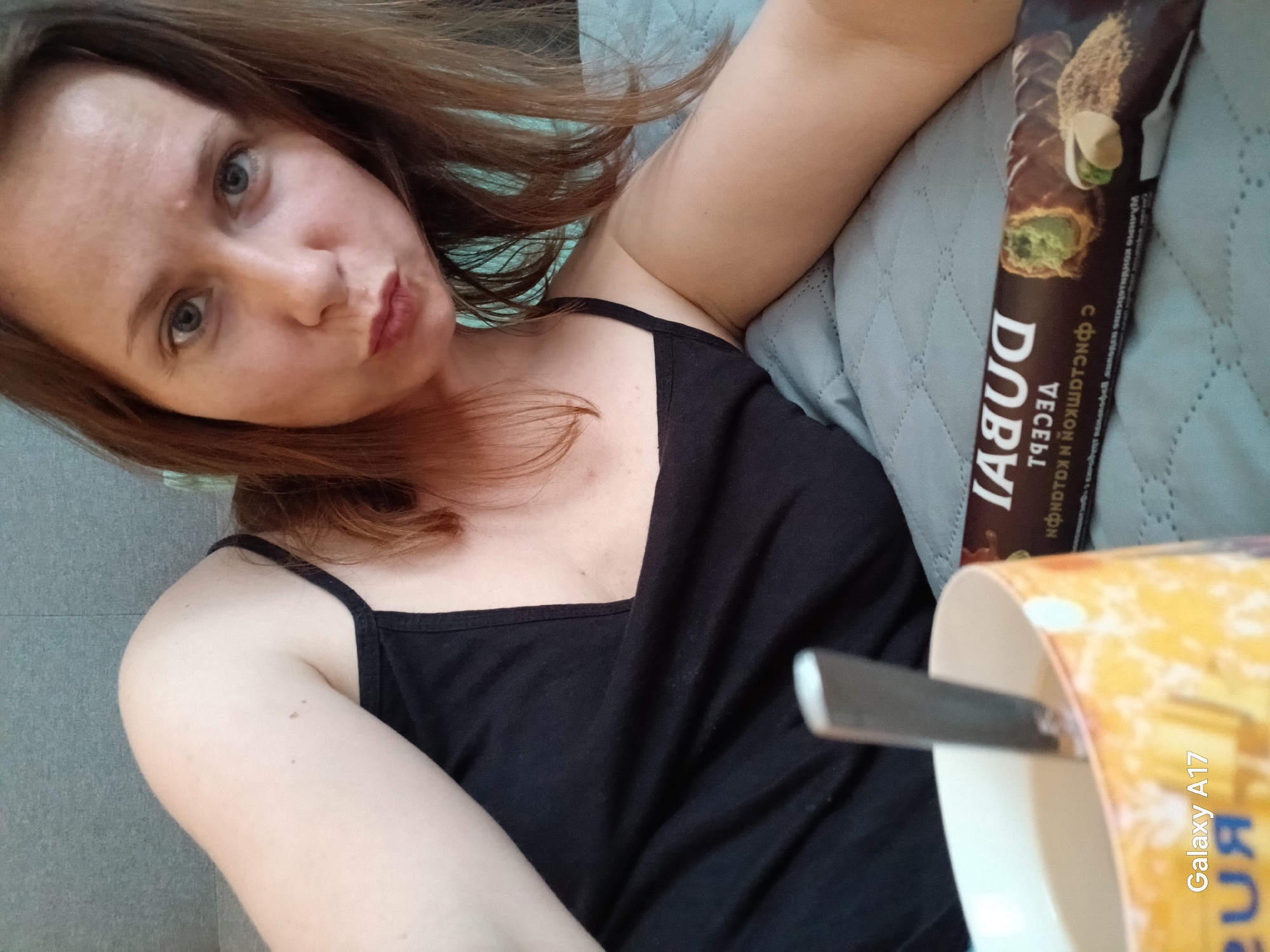 A selfie taken in Yekaterinburg, Russia, shows a woman with light skin and reddish-brown hair, wearing a black tank top. She is looking at the camera with a slight pout. In the background, there is a teal quilted fabric, possibly a blanket or cushion. To the right of the woman, there is a chocolate bar with pistachio filling, with the word "ДУБАЙ" in Russian printed on the wrapper. A mug is also visible with a spoon sticking out of it. "Galaxy A17" is printed in the bottom right corner of the image.