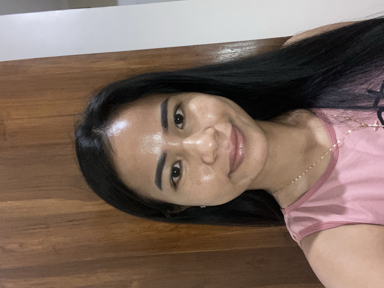 A close-up, portrait-style photograph, rotated 90 degrees clockwise, captures a young adult woman smiling gently. She has long, straight, dark hair that cascades over her right shoulder and down her back. Her complexion is smooth, with a subtle shine visible on her forehead and nose, suggesting a natural or dewy look. She has neatly groomed dark eyebrows, dark eyes, and full lips with a light pink or nude lip color.
The woman is wearing a light pink sleeveless top, possibly a tank top, with a darker pink trim around the neckline. A delicate gold-colored chain necklace with a small pendant is visible around her neck.
The background predominantly features a warm-toned wooden surface, likely a wall or paneling, running vertically behind her. A narrow strip of a plain white surface is visible at the top edge of the image (which would be the top right if the image were correctly oriented). The lighting appears soft and even, highlighting her features. The overall mood conveyed is pleasant and content. This indoor setting is located in North Kuta, Indonesia. No visible text is present in the image.