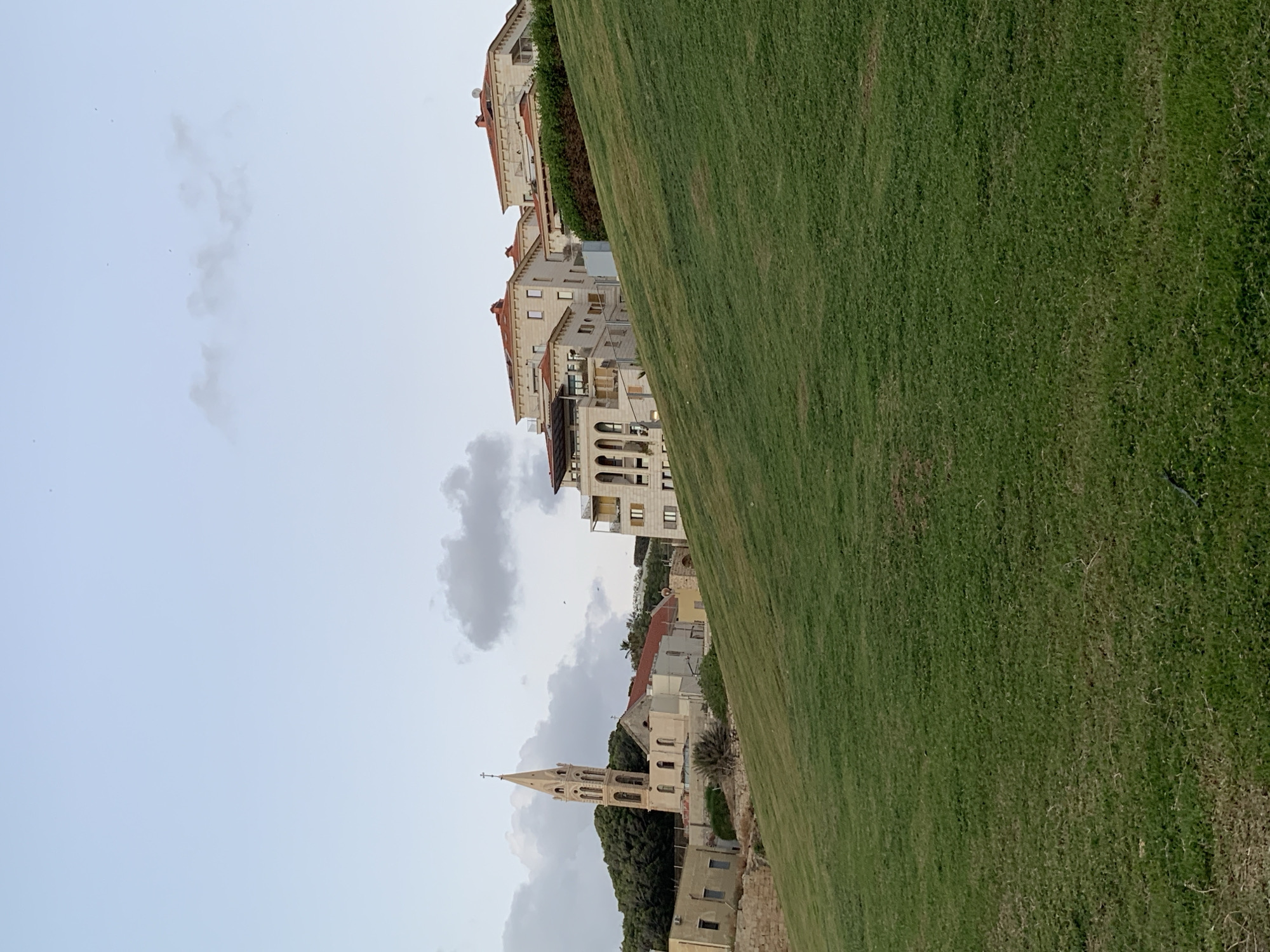The image depicts a view of the city of Tel Aviv, Israel. The scene includes a sloping green lawn in the foreground, with buildings and structures visible in the midground and background. Among the buildings is the St. Peter's Church, characterized by its tall, pointed bell tower. The sky above the buildings is partly cloudy. The photograph is composed with a tilted perspective.