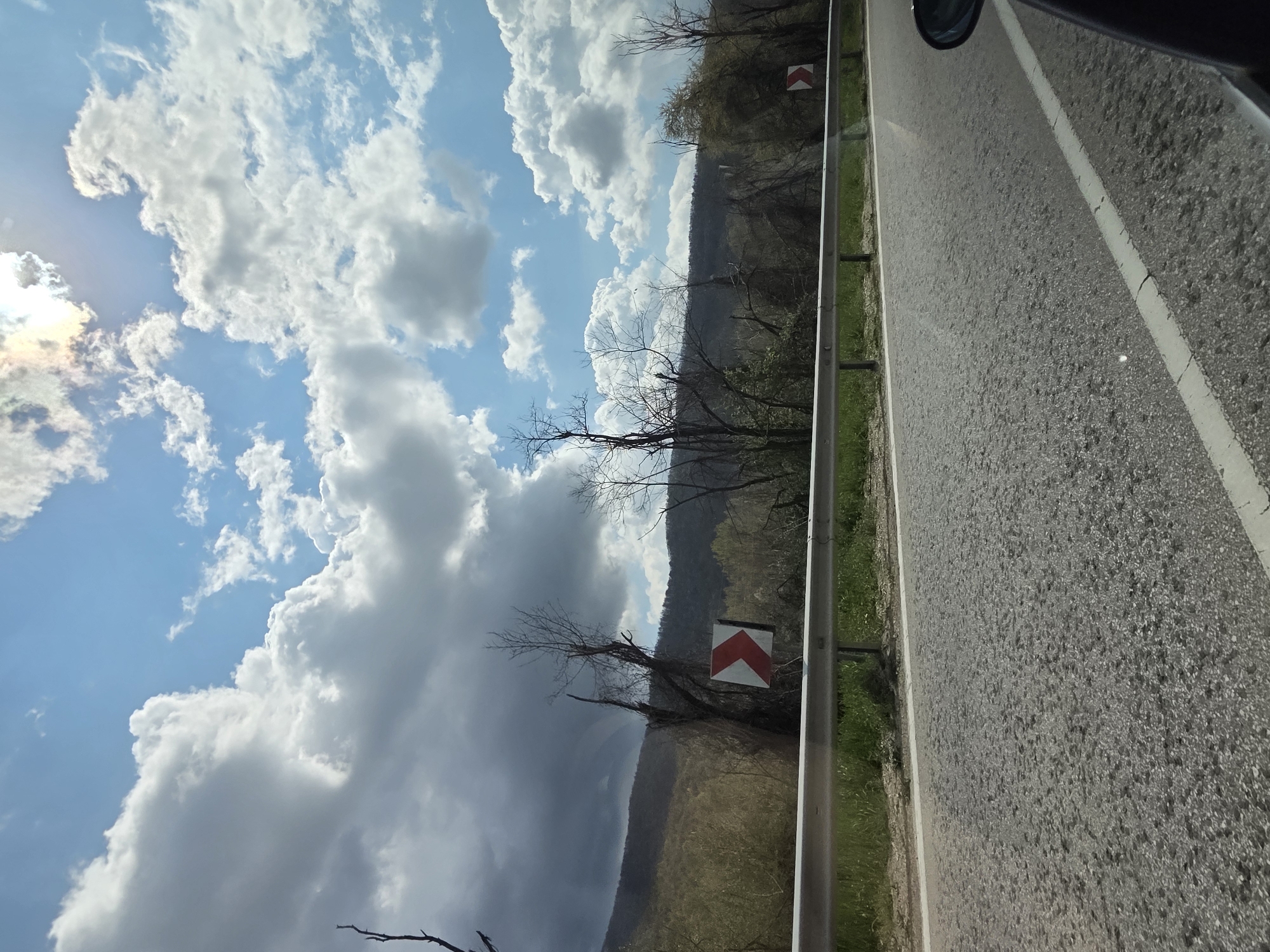 The image shows a road next to a metal guardrail. The sky is partially cloudy, with blue sky visible between the clouds. There are trees and a hillside visible on the other side of the guardrail. A road sign with a chevron is visible. The location appears to be in a rural area. Based on the user's description, it is located in Leshnitsa, Bulgaria.
