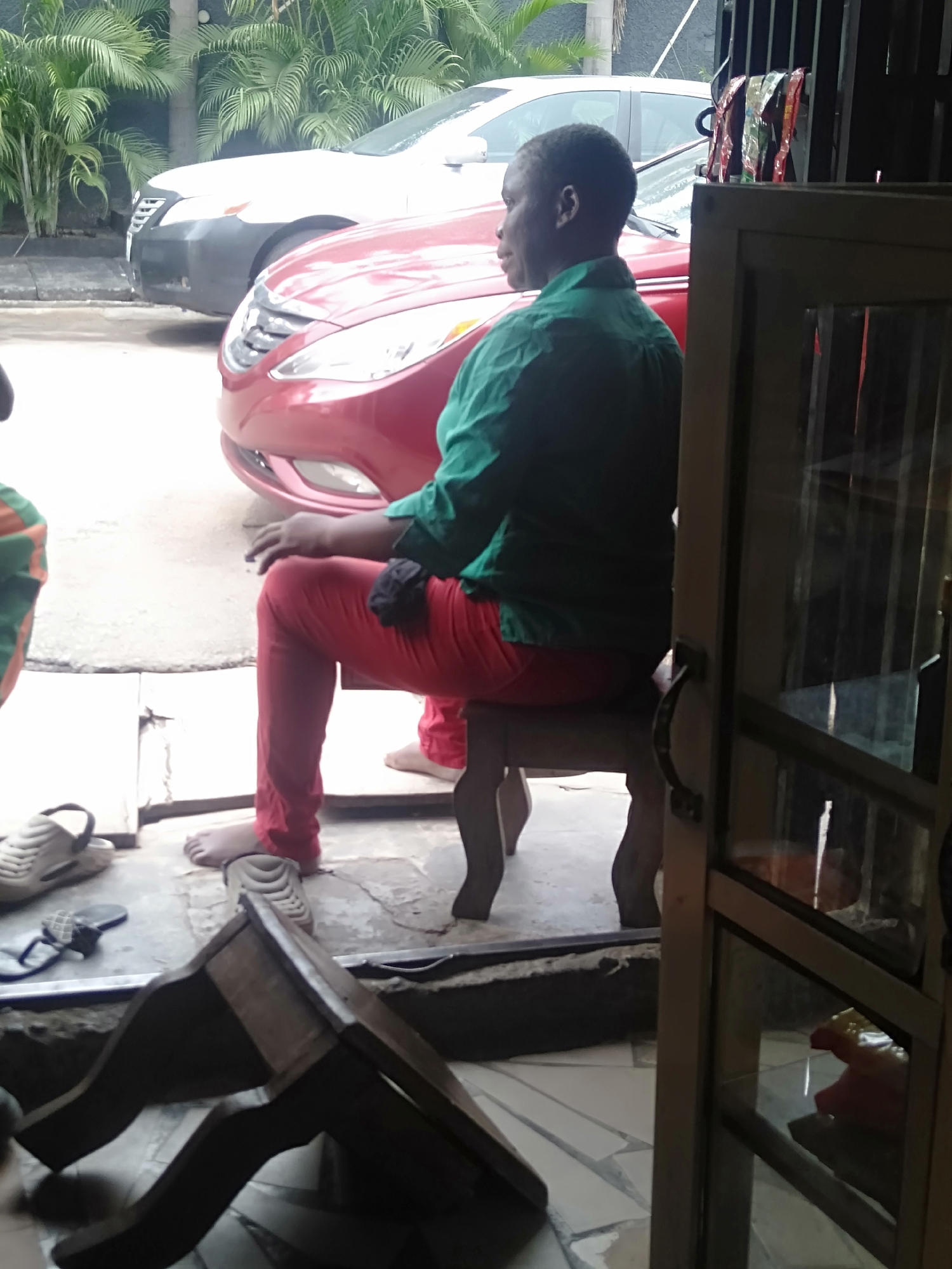 An image taken in Shangisha, Nigeria, depicts a man sitting on a wooden stool outside a building. The man wears a green, long-sleeved shirt and red pants. He is barefoot. In the background are two parked cars, one red and one white, along with some foliage. A second wooden stool is overturned on the floor near various items of footwear. The image is partially obscured by a door frame.