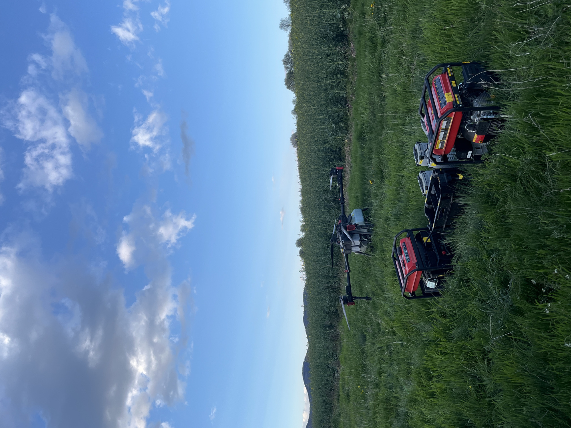 The image shows a grassy field under a blue sky with scattered clouds. Several objects are visible in the field: four portable generators, red and black in color, and two unmanned aerial vehicles (UAVs), also known as drones, with extended arms. The background features a line of dense vegetation, possibly bushes or trees. The location appears to be outdoors and rural, but I cannot confirm the exact location in Varbeshnitsa, Bulgaria without additional context or landmarks.