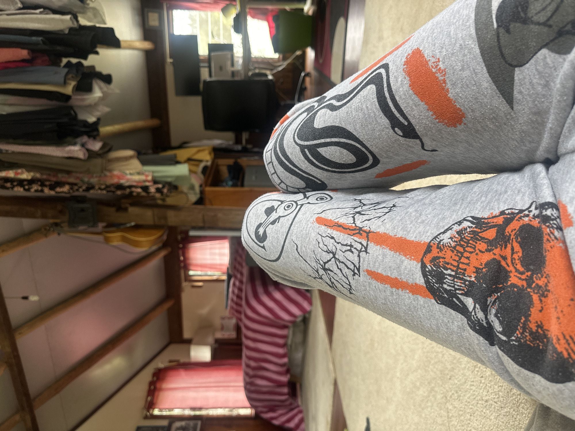A first-person perspective view of a person's legs, clad in grey sweatpants adorned with a skull and a stylized graphic design, dominates the foreground. The sweatpants feature a black and orange skull on the left leg and a black, swirling design with orange accents on the right leg. The background reveals a cluttered room with a sloped ceiling, exposed wooden beams, and hanging clothes. A bed with a pink and white striped comforter is visible in the mid-ground, with red curtains framing a window. Further in the background, a desk with a computer monitor, a black office chair, and shelves filled with various items are present. The overall impression is that of a lived-in, informal living space. The location is not specified in Thora, Australia.