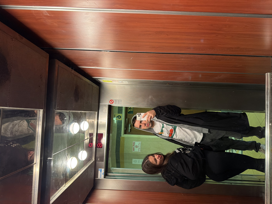 The image depicts an interior view of an elevator, occupied by two individuals. The right wall of the elevator is constructed from reddish-brown wood paneling separated by vertical metallic strips. The left wall and the elevator doors are made of stainless steel. The elevator ceiling features a central mirrored panel reflecting the interior, bordered by perforated metal panels. Four spherical white light fixtures are mounted on the mirrored ceiling. A red digital display above the elevator door shows the letter "E," indicating the current floor.
A male individual, positioned towards the left of the frame, is taking a mirror selfie using a white smartphone held in his right hand. He wears a grey hooded sweatshirt underneath a black long jacket or coat, and dark grey pants. His expression is neutral as he looks into the phone's camera. To his immediate left, a female individual stands with her back partially turned, glancing over her right shoulder towards the male. Her lips are pursed in a pout, and she has long, dark hair. She is dressed in a black hooded sweatshirt with white markings on the back, and black form-fitting pants.
Through the open elevator doors, a hallway is visible, featuring light green walls with a horizontal yellow stripe and white door frames. Signs with Hebrew text are reflected on the elevator door from the hallway. A yellow sign on the door frame reads "שבת חובה" (Shabbat obligatory/mandatory), and a white sign on the green wall below it states "אסור לצאת" (Forbidden to exit). These Hebrew signs identify the location as a building in Israel, likely an urban residential setting such as Tel-Aviv, which commonly features Shabbat elevators.