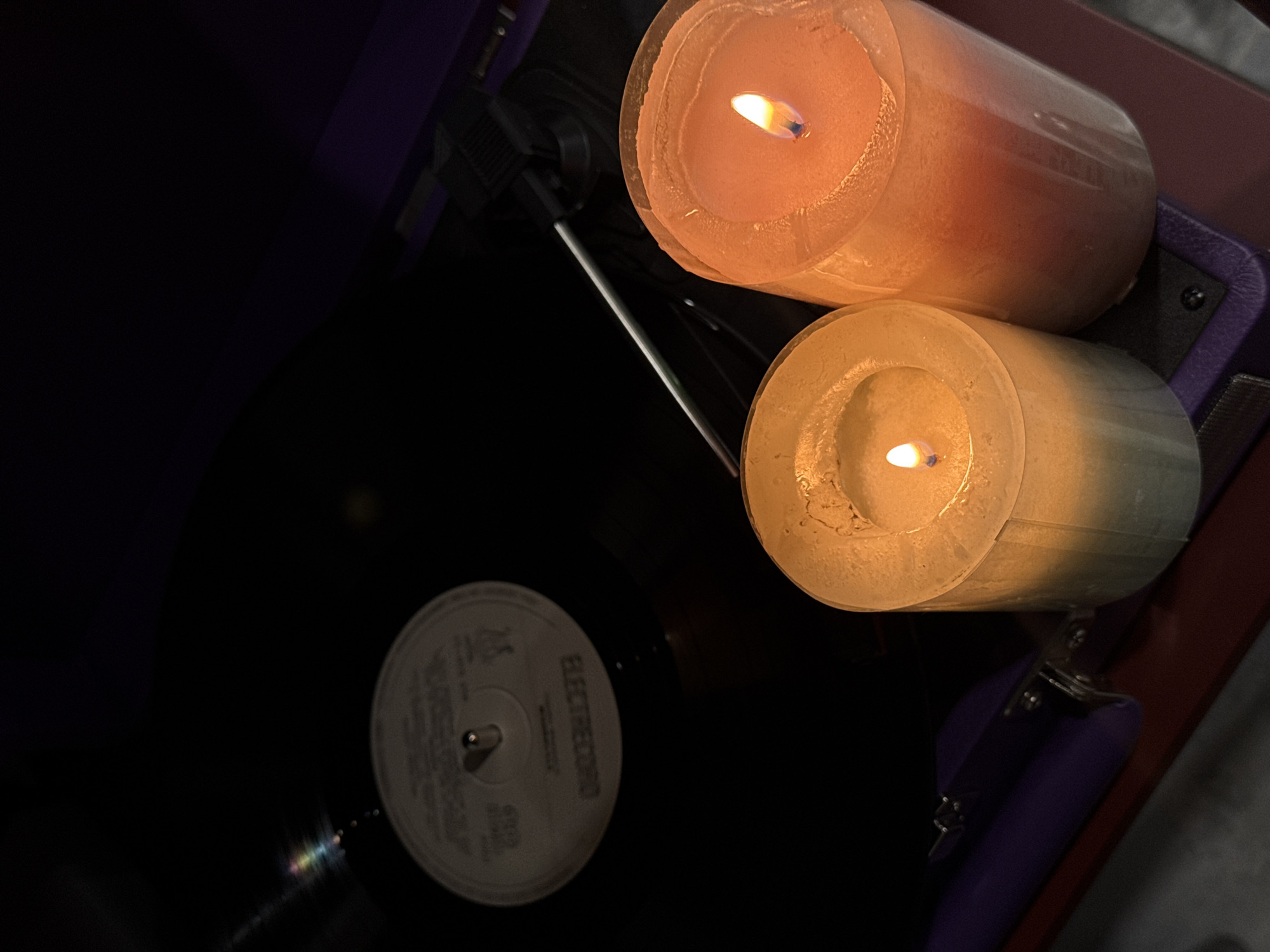The image shows a close-up, overhead view of two lit candles, a vinyl record, and part of a record player. The candles are cylindrical and sit next to each other; the top candle is peach-colored, while the bottom candle is white. Both candles have visible flames. Below the candles, a vinyl record is partially visible, with its label facing upwards and slightly obscured. The label contains text. To the left of the candles and record is a record player, with part of the tone arm. The scene is dimly lit, with the candles providing the primary source of light. The image was not taken in Yerevan, Armenia.