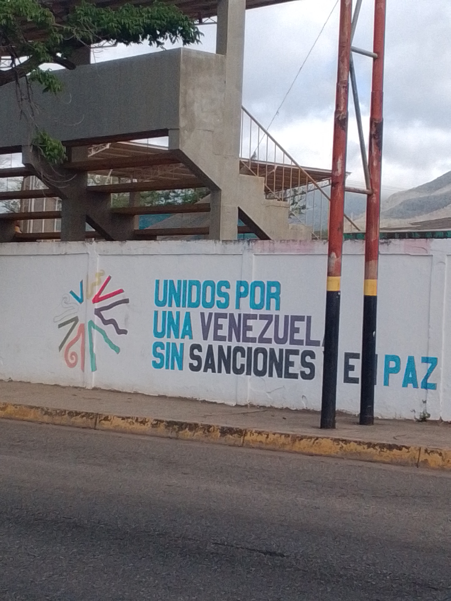 A mural is painted on a white concrete wall in Altagracia, Venezuela. The mural features a graphic of a sun-like symbol on the left, with colorful, abstract lines radiating outwards. To the right of the symbol, the text "UNIDOS POR UNA VENEZUELA SIN SANCIONES E PAZ" is painted in large, bold letters. The words "UNIDOS POR UNA VENEZUELA" are in a bright blue color, while "SIN SANCIONES" is in dark gray. The final word, "PAZ," is in the same bright blue as the first part of the text. The mural is located outdoors, with a street and sidewalk in the foreground. In the background, there is a concrete structure with stairs and what appears to be a canopy. Two red poles with black and yellow accents stand in front of the wall, partially obscuring the text. The sky is overcast, suggesting a cloudy day. The overall scene depicts a message of unity and peace for Venezuela, specifically referencing the absence of sanctions.