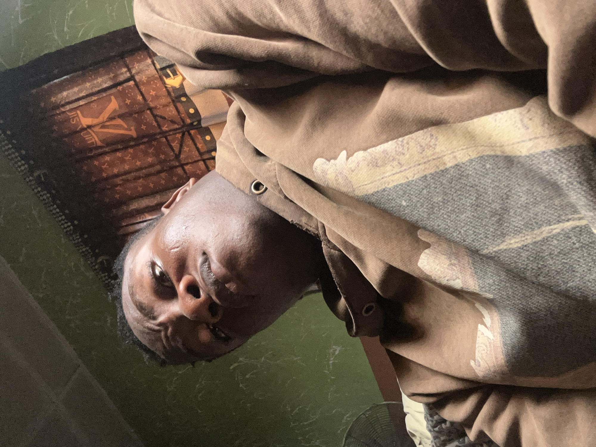 A close-up, low-angle shot shows a person with dark skin, wearing a brown hoodie, possibly indoors. Part of a dark-colored object with what appears to be a "LV" pattern is visible behind the person. The location is assumed to be Ikorodu, Nigeria. Other objects visible are a fan in the bottom corner and a dark-colored wall in the background. The image quality is somewhat grainy.