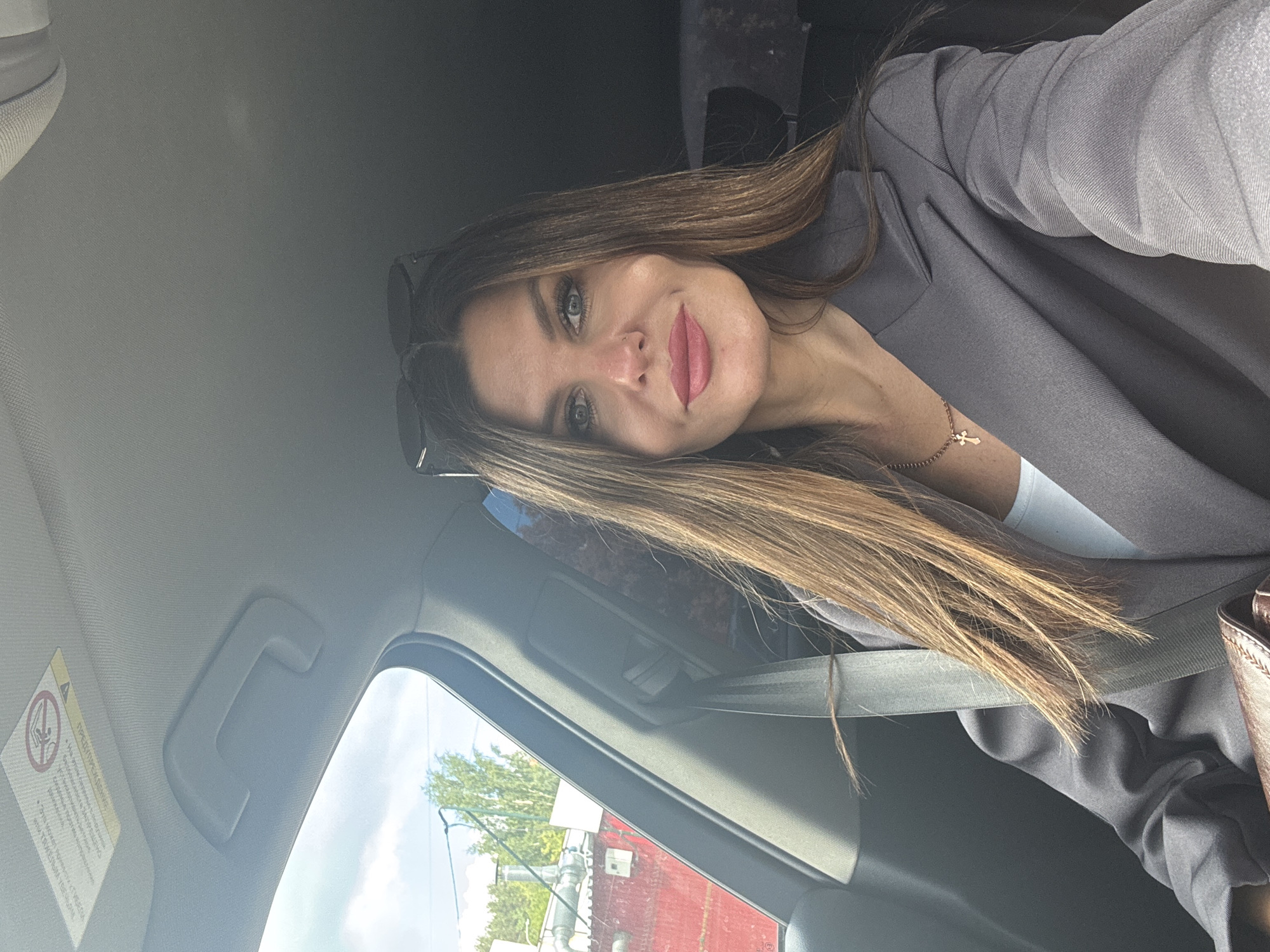 A selfie taken by a woman inside a car. The woman is wearing a gray blazer, a white top, and a necklace with a cross pendant. Sunglasses are perched on top of her head. The car's interior is visible, including the seatbelt, dashboard, and roof. Through the window, one can see part of a building with a red wall and a metal structure, along with some trees. While the specific location is not determinable from the image, the city of Chaikovsky, Russia, is mentioned.