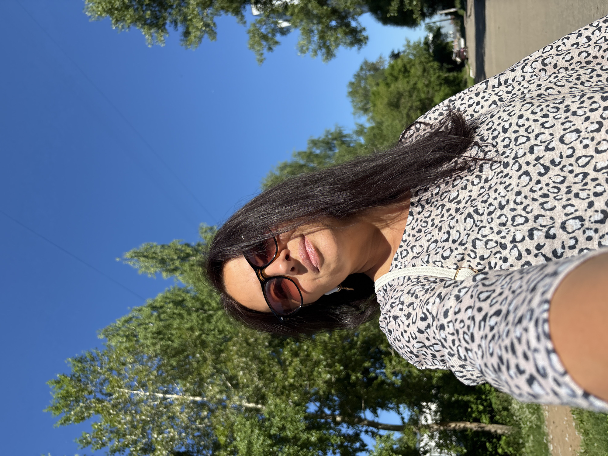 A medium shot captures a woman with long, dark hair wearing large, dark-rimmed sunglasses and a light-colored top with a black leopard print. She is holding a camera or phone out to take a selfie. The woman stands on a paved path, likely a sidewalk or street, with trees lining both sides. The sky is a clear, bright blue, and the sun is casting a strong light, creating distinct shadows and highlights. In the background, there is a suggestion of a residential area with houses and a road. The image appears to have been taken in Chaikovsky, Russia, on a sunny day.