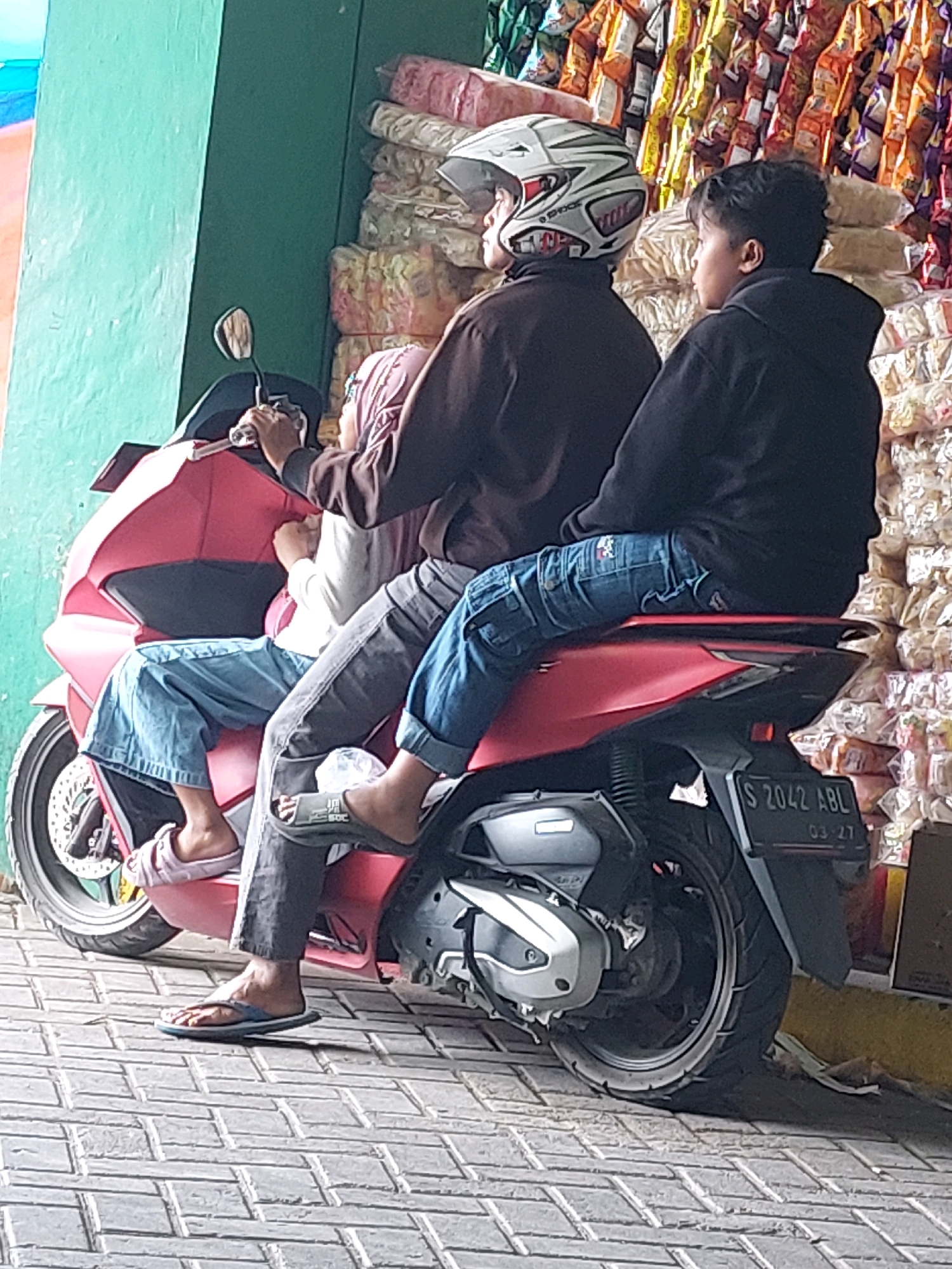 Three individuals, likely a family, are seated on a red and black scooter in what appears to be a commercial area in Sumuragung, Indonesia. The person in the front, wearing a white and silver helmet, is likely the driver. They are positioned behind a handlebar and mirror, indicating control of the vehicle. The middle person, wearing a pink headscarf and white shirt, is seated behind the driver, with their legs positioned on the scooter's front body. The third person, a young male wearing a black hoodie, is seated at the rear of the scooter, his legs extended. The scooter is parked on a brick-patterned sidewalk, adjacent to a wall painted in a muted green color on the left. To the right of the scooter, stacks of brightly colored packaged goods, possibly snacks or food items, are displayed, suggesting a market or shop setting. The license plate on the rear of the scooter is visible and reads "S 2042 ABL 03-27". The lighting suggests it is daytime.