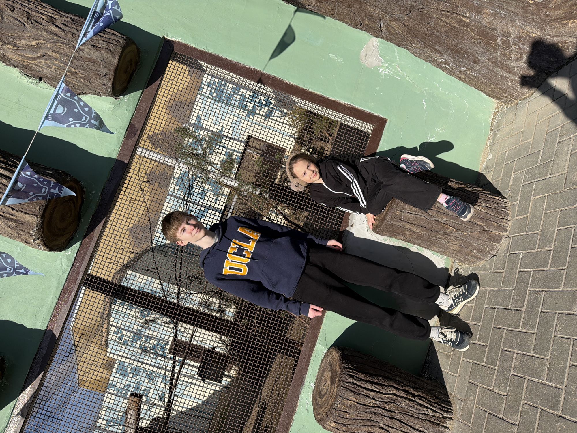Two people are lying on artificial logs inside an enclosure with a metal grid roof. The person on the left is wearing a dark blue UCLA hooded sweatshirt and black pants. The person on the right is wearing a black tracksuit with white stripes. The location appears to be an outdoor zoo or animal park, identifiable as the Perm Zoo in Perm, Russia, due to the architectural style of the enclosure and adjacent brick paving pattern. The environment consists of artificial logs and a green painted wall. Bunting or flags are visible to the left.