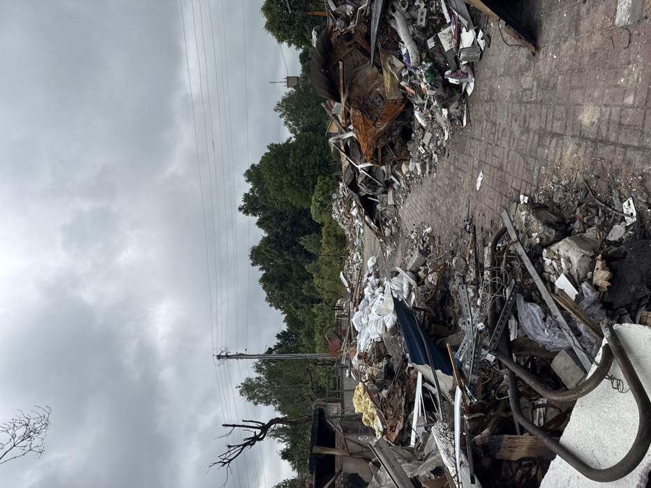 A landscape of destruction. A path of red bricks barely visible amidst heaps of debris, burnt metal, and concrete fragments. Piles of white bags suggest remnants of possessions. Trees stand in the background under a gloomy, overcast sky, juxtaposing nature's permanence with the scene's devastation. Electrical wires stretch across the sky, connecting to poles that look as if they might be the next casualties of whatever occurred.