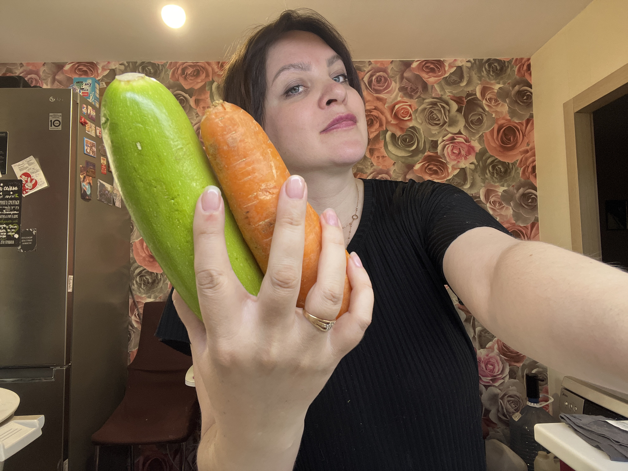 Here is a detailed description of the image:
A woman is holding a zucchini and a carrot in her left hand, posing for a selfie. She has short, dark hair, fair skin, and is wearing a black shirt. She has a ring on her ring finger.
The background includes a refrigerator with magnets and notes on it, and a floral wallpaper. A countertop and a portion of a kitchen appliance are also visible on the right side of the frame.
This image was taken in Noviy, Russia.