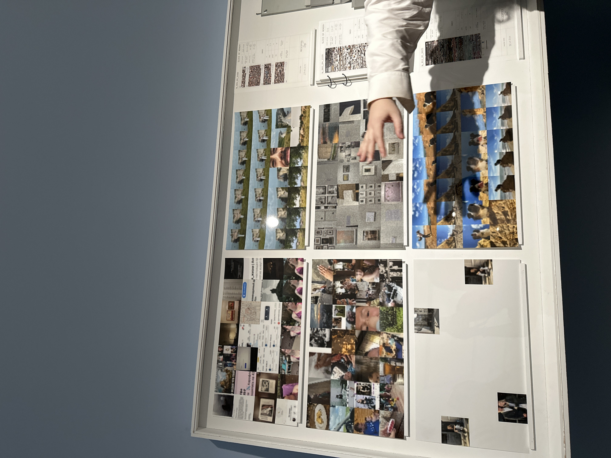 The image shows a framed display hanging on a light blue wall. The display is divided into several sections containing various images, seemingly arranged as a collage. Some sections contain tiled arrangements of smaller images, while others have larger, individual images. There are also documents with printed text and tables, possibly detailing information related to the images. A hand with a white sleeve is partially visible, appearing to interact with the display. The environment is simple, with a single-colored wall as the backdrop, focusing attention on the displayed images and documents. The OCR'd text indicates references to "SLICE OF MEMORY" and a "RECEIPTED" document. The location is unknown, and there is no clear indication this image was taken in Moscow, Russia.
