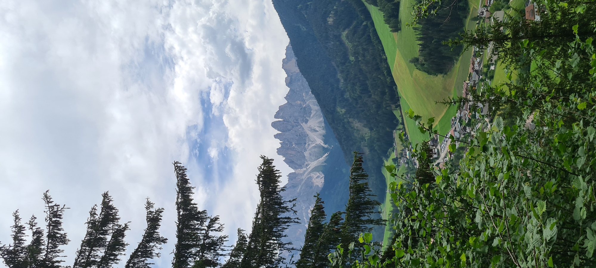 The image is a landscape photograph captured from a vantage point overlooking the village of St. Magdalena (Santa Maddalena), Italy. In the foreground, dark green foliage of trees and bushes partially obscures the view, with pine branches reaching in from the upper left. The midground reveals a picturesque valley dotted with the red-tiled roofs of houses belonging to St. Magdalena, nestled amongst vibrant green meadows and darker green forests. A road with parked cars is visible within the village. The background is dominated by a dramatic mountain range with jagged, rocky peaks, some of which are dusted with snow. The sky above is a mix of white and gray clouds, with patches of blue sky peeking through. The overall scene depicts a serene alpine village with imposing mountains as a backdrop.