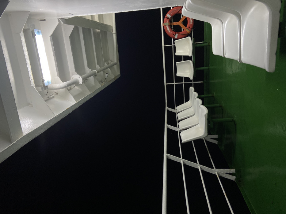 Captured at night, the scene depicts a ship's deck. On the left, a white structure houses a bright fluorescent light, revealing a ribbed interior. To the right, a green wall is adorned with a row of white chairs secured by a metal railing. A life preserver hangs above two of the chairs. The dark sky occupies the space between the deck structures.