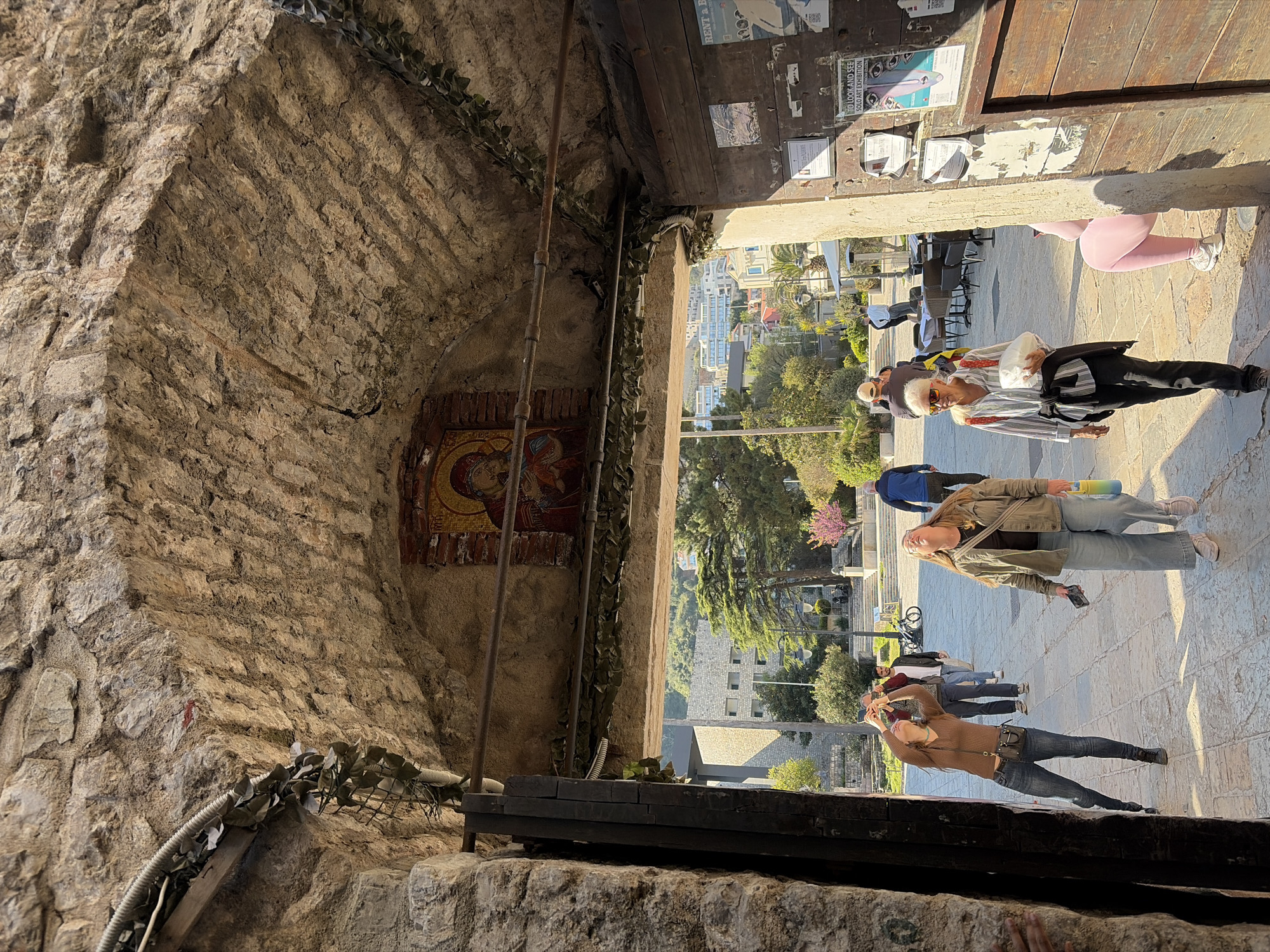 A street view in Budva, Montenegro, with multiple people visible. One person is in pink leggings. Two people are taking photos. A white-haired person is walking. The buildings are visible in the distance. The photo is taken from within a stone structure, possibly a tunnel or archway, with a religious image on one wall and advertisements on the other.