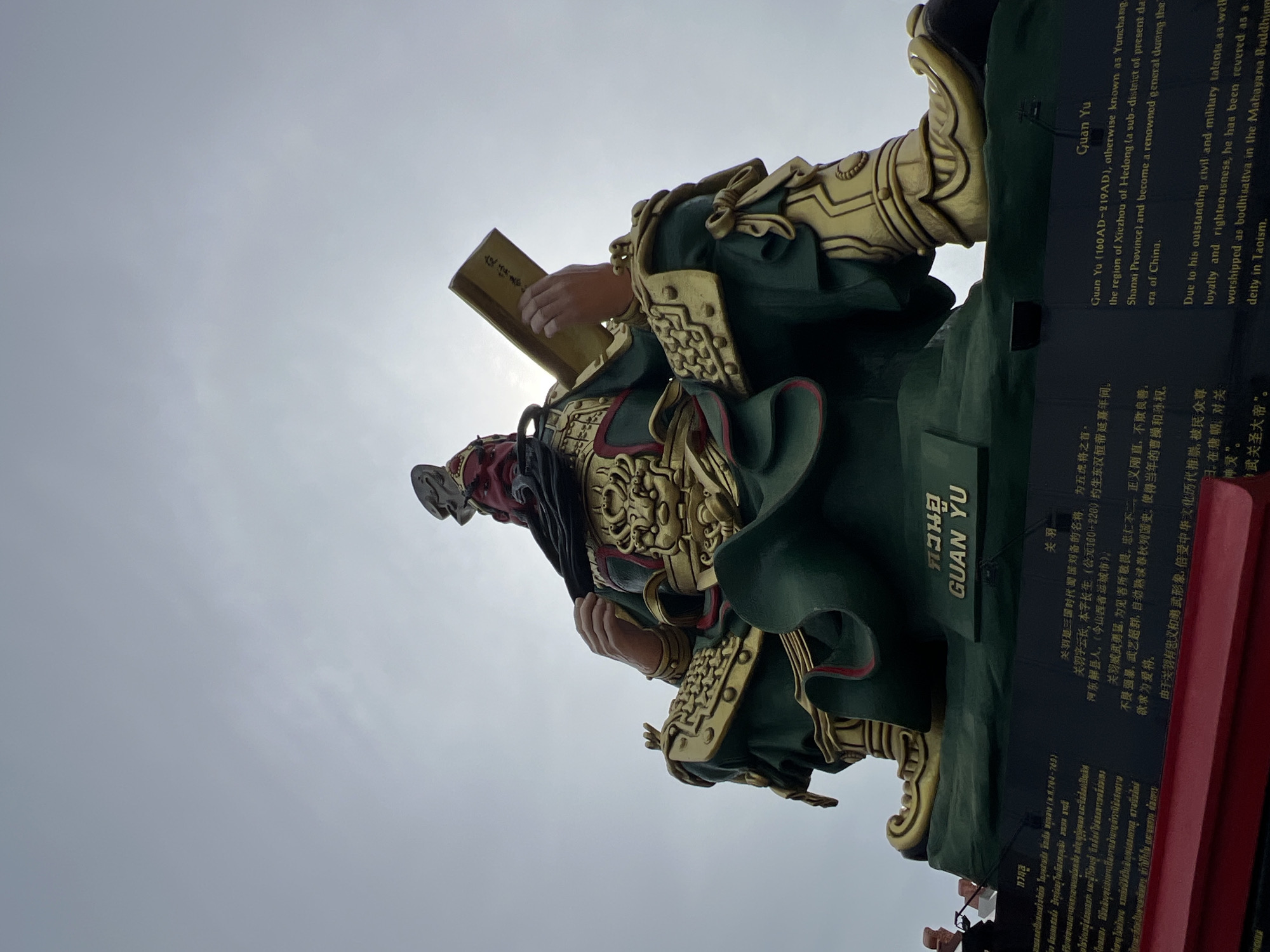 A low-angle, wide shot captures a colossal statue of Guan Yu, a deified Chinese general. The statue is predominantly green and gold, with a red face and beard, and it sits cross-legged, holding a scroll. The statue is placed on a dark base, which features text in both Thai and English. The English text identifies the subject as "Guan Yu (160AD-219AD), otherwise known as Yumachang, from the region of Xiezhou of Hedong (a sub-district of present-day Shanxi Province) and become a renowned general during the era of China." It also states that "Due to his outstanding civil and military talents as well as loyalty and righteousness, he has been revered as a deity in Taoism." The statue is located in Baan Hua Thanon, Thailand, and is set against an overcast, pale gray sky.