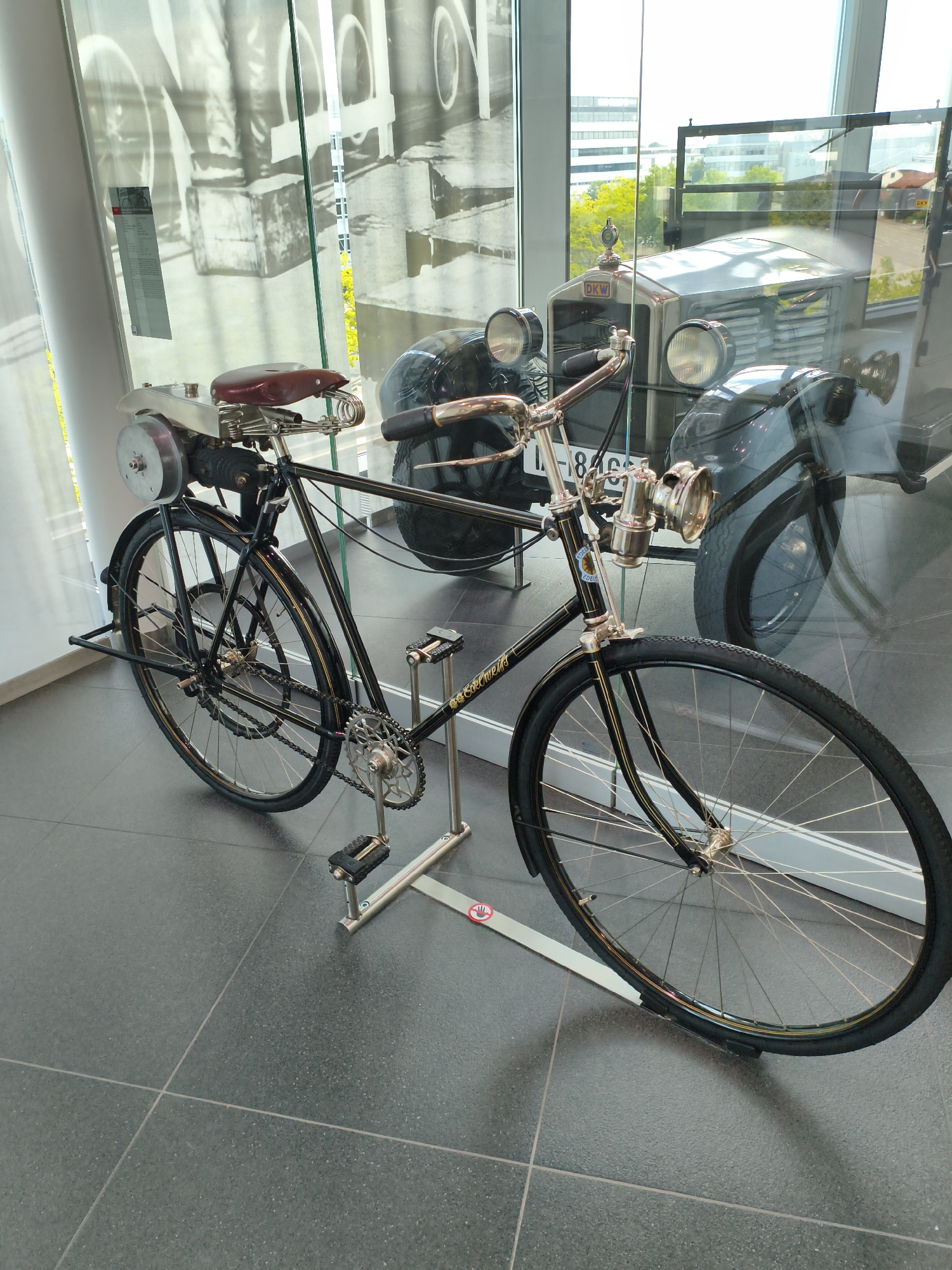 An antique motorcycle is displayed on a stand in a museum. The motorcycle has a black frame, a brown leather seat, and a large headlight. It is positioned on a dark gray tiled floor, with a reflective glass wall behind it. Through the glass, a vintage car, also on display, can be seen, along with the cityscape of Ingolstadt, Germany, visible in the background. The car is silver with a black grille and two round headlights. The motorcycle has a chain-driven rear wheel and a kickstand. The scene appears to be indoors, likely in a museum or exhibition space.