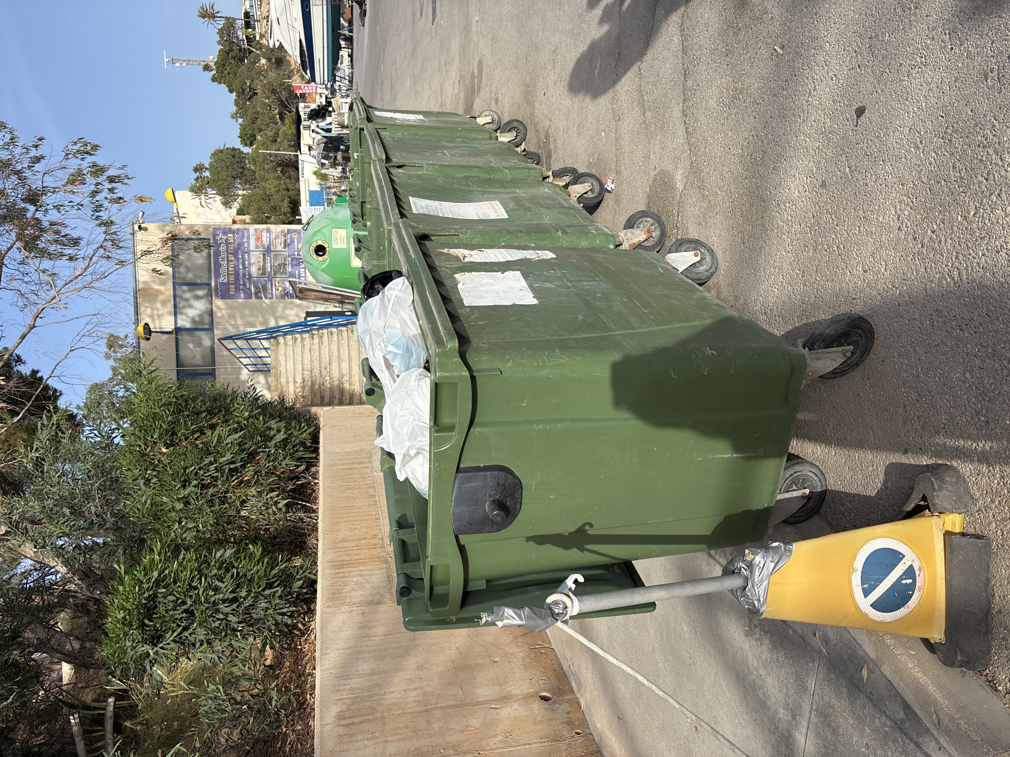 A line of large, green, wheeled trash receptacles is parked on an asphalt surface. The receptacle closest to the viewer is partially filled with white plastic bags. A metal pole with a yellow traffic cone at its base, secured with silver tape, is positioned in front of this trash receptacle. The cone features a blue and white circular symbol indicating a prohibition. Behind the trash receptacles, a concrete wall is visible, topped with dense green foliage. Further back, a building with a blue-framed window and a sign that reads "Stella Maris FOR THE LOVE OF THE SEA" is present. Blue stairs lead up to the building entrance. In the background, a clear blue sky is visible, along with trees and several boats docked. The scene appears to be located in a coastal or marina area.