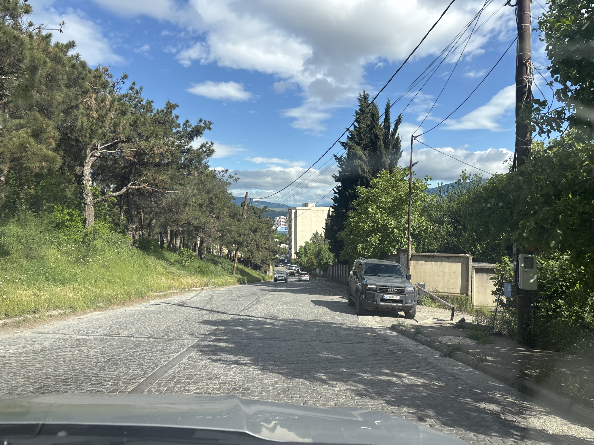 Captured from inside a vehicle, the image presents a street scene in Tbilisi, Georgia, characterized by a cobblestone road leading uphill. On the right side of the road, a dark gray Toyota SUV is parked, license plate visible. A concrete wall and dense vegetation line the roadside. To the left, a grassy area and a row of pine trees border the street. In the distance, several cars ascend the hill, passing in front of a building complex. Overhead, power lines stretch across a blue sky dotted with cumulus clouds. A utility pole stands on the right side of the frame. The vehicle's dashboard is partially visible at the bottom of the image, suggesting the viewpoint is from the driver's seat.