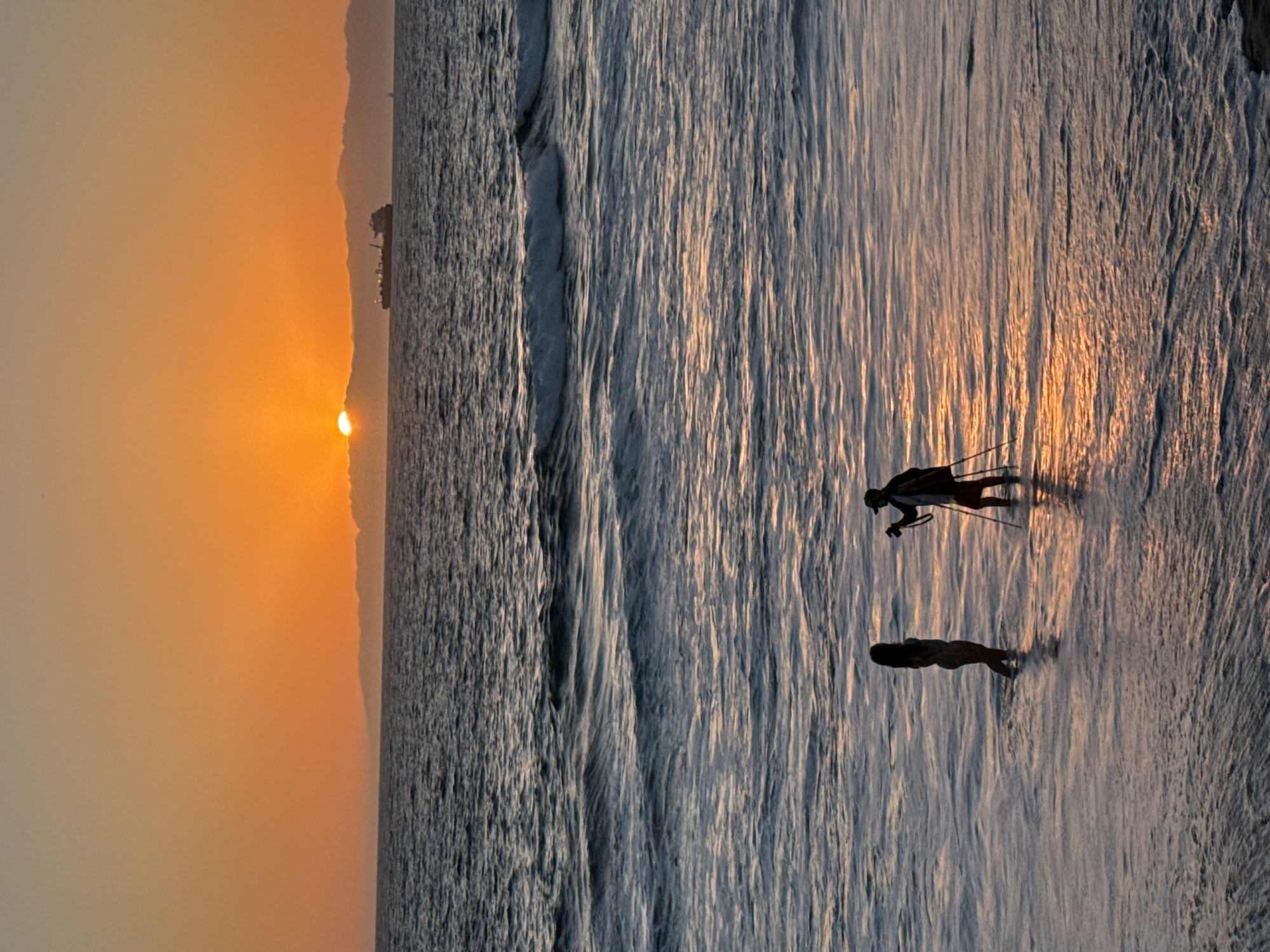 In Manhattan Beach, California, at dusk, two people are in the shallow ocean water near the shore. A person on the left, silhouetted against the orange sky, stands facing right, appearing to be looking at the person on the right. The person on the right, also silhouetted, is facing the person on the left and is holding a camera on a tripod. The sun is low on the horizon, casting a warm glow on the water and sky. A cargo ship is visible in the distance on the ocean. The waves are gentle, and the water reflects the colors of the sunset. The beach sand is submerged in the water.