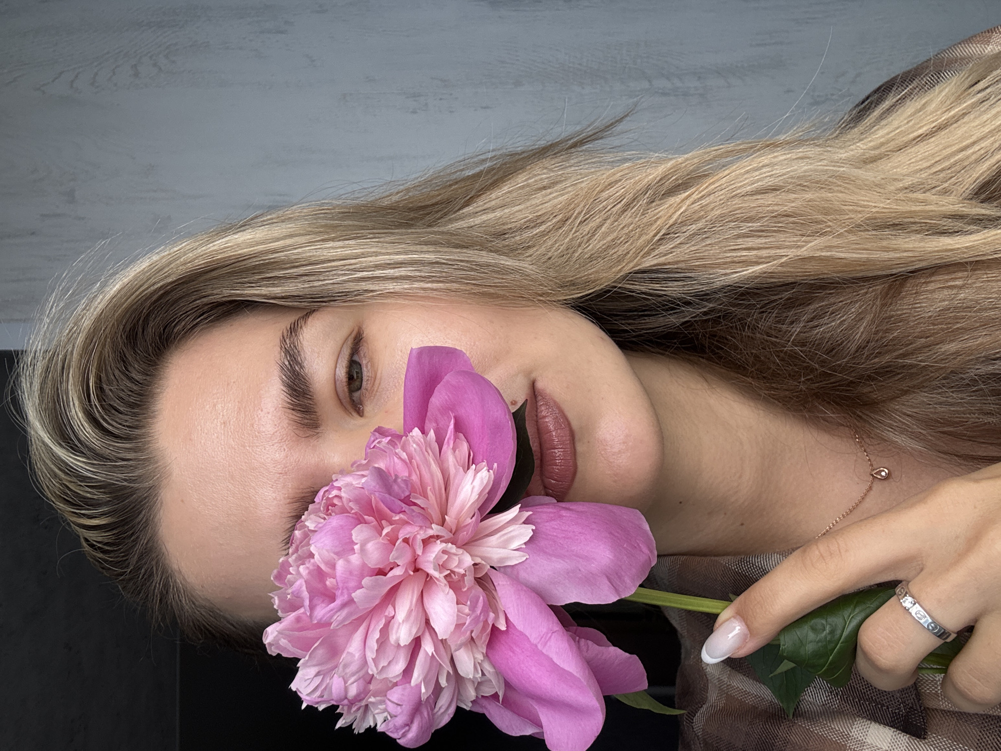 A young adult female with long, wavy blonde hair and visible dark roots poses with a large pink peony flower in the city of Unknown, Unknown. The flower is held by her right hand, partially obscuring the left side of her face, from the forehead to the chin. Her visible right eye is green, and her eyebrows are thick and dark. Her lips are painted with a dark nude or brownish lipstick. She wears a brown plaid or checkered garment. On her right hand, her nails are manicured with white tips. A silver-toned ring with three square-set clear stones is on her right ring finger. A delicate rose gold-toned necklace with a small teardrop-shaped pendant containing a single clear stone is visible on her neck. The background to her left is a solid dark gray or black wall, while the background to her right is a light gray, textured wall with vertical lines, suggesting wood paneling or concrete.