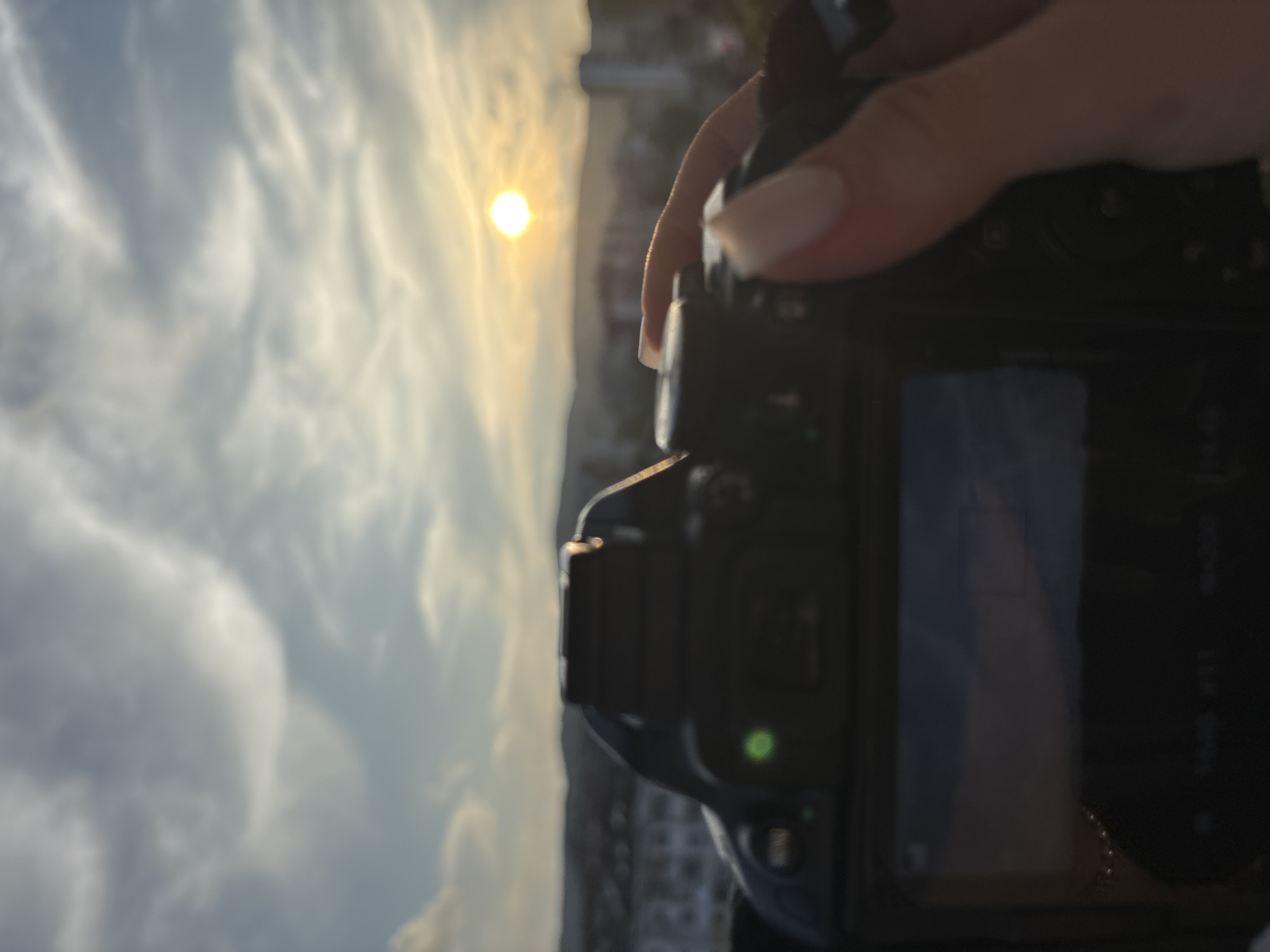 Here is a description of the image:
The image shows a person holding a black digital camera in their right hand. The camera is out of focus. In the background, there is a view of the sky with clouds and the sun. The image is taken from a high vantage point, possibly a rooftop or tall building. The buildings below are blurred, and the location is not identifiable. The person is holding the camera to capture a photo of the sunset.