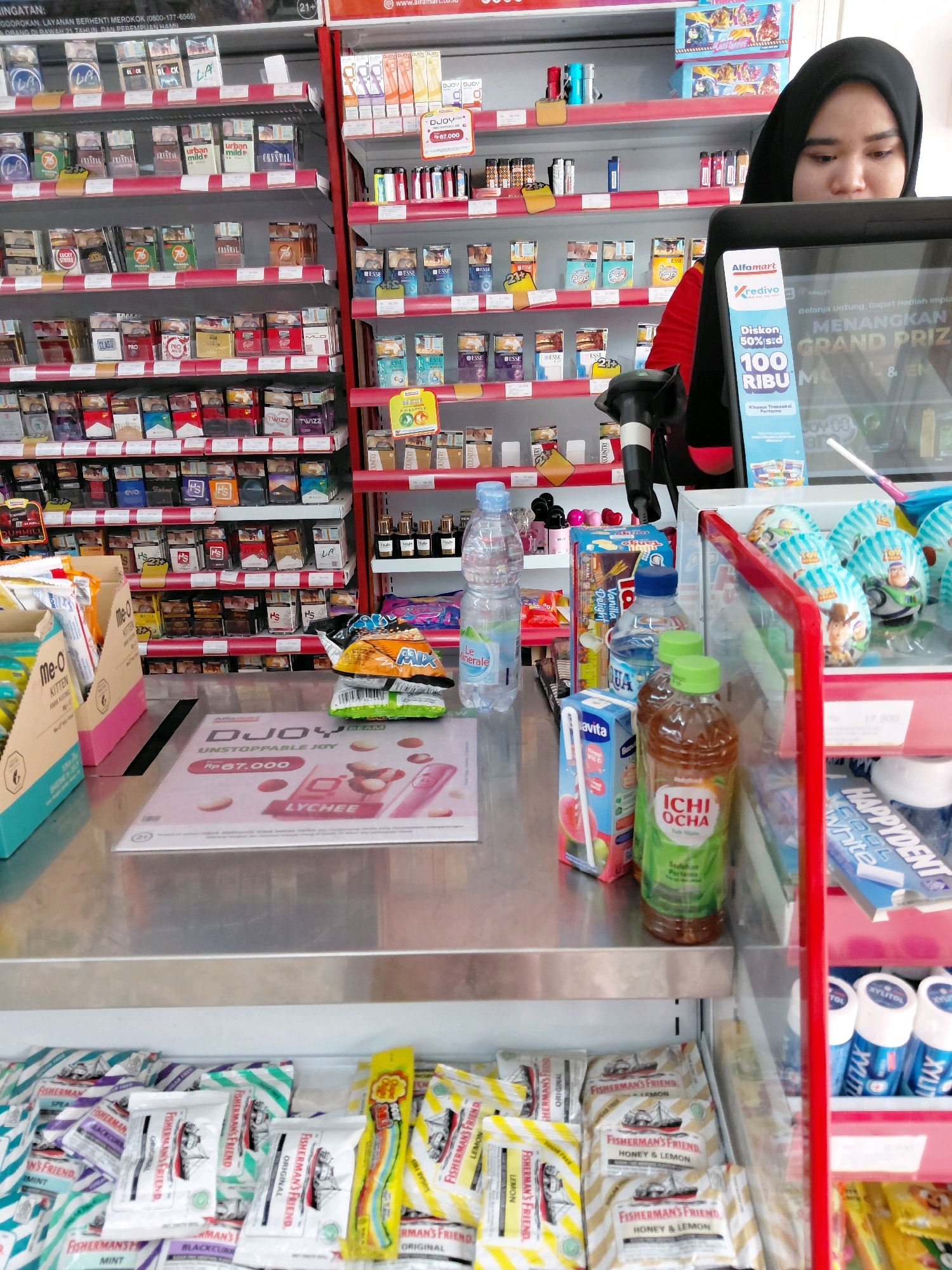The image shows a convenience store counter in Amin Jaya, Indonesia. A cashier, a woman wearing a black hijab, is partially visible behind the counter, looking at a monitor. On the counter, there are several items including a stack of chips, a bottle of water, and a paper flyer promoting a "DJOYO UNSTOPPABLE JOY" product. Behind the cashier, shelves are stocked with various products, including cigarettes, snacks, and drinks. The overall environment appears to be a typical retail setting.