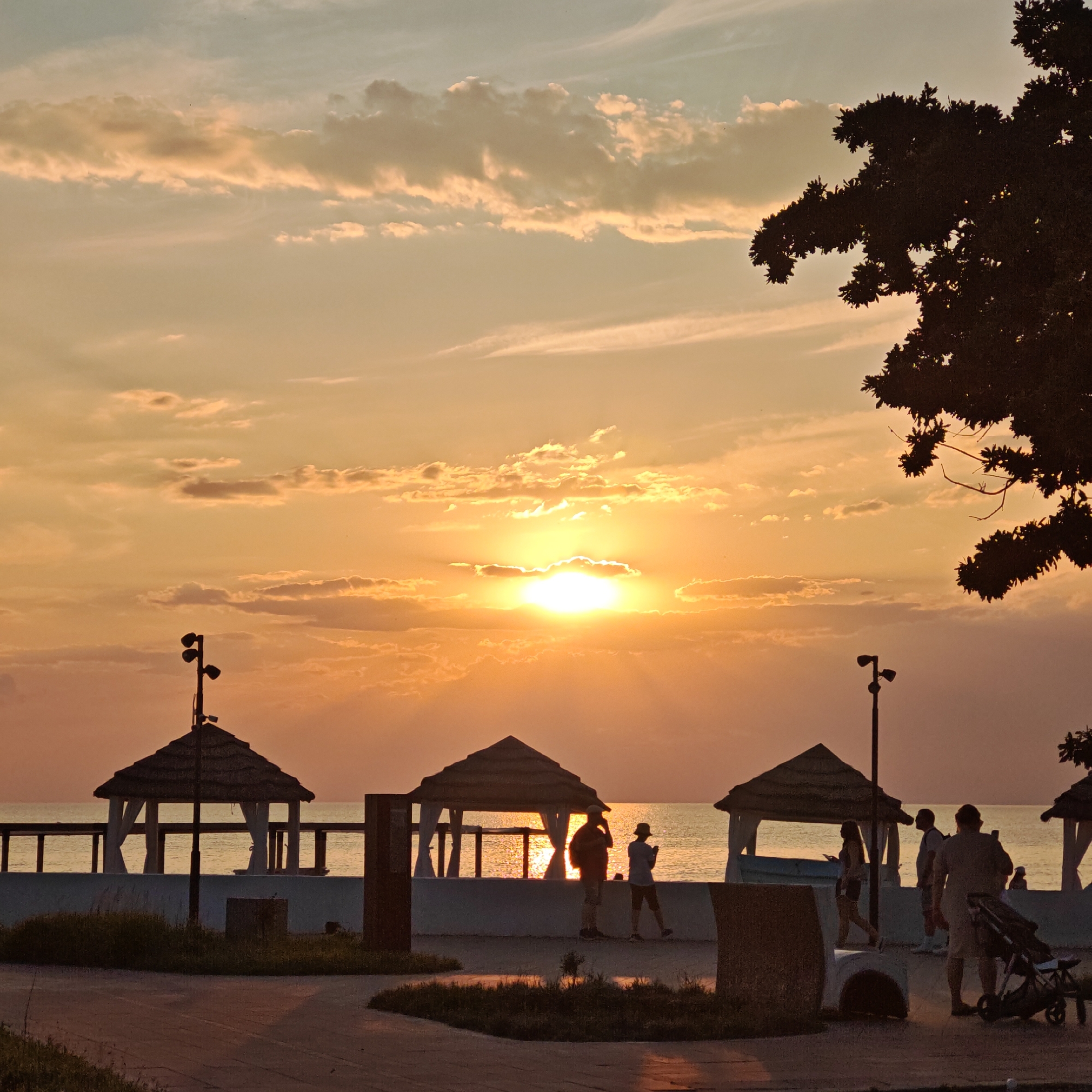 The image depicts a serene sunset scene by the sea. The sky is ablaze with warm hues of orange and yellow, with scattered clouds catching the golden light. The sun is low on the horizon, casting a shimmering reflection on the calm water.
In the foreground, silhouetted figures of people are visible, enjoying the evening ambiance. Some are walking along a paved path, while others are gathered near beachside gazebos. The gazebos, with their thatched roofs and white curtains, suggest a resort or recreational area. There's a lamppost near one of the gazebos, and a stroller is parked near a group of people. The overall setting appears to be a promenade or boardwalk overlooking the beach.
The time of day is clearly sunset, indicated by the low angle of the sun and the warm, diffused light. The weather seems pleasant and calm, with no strong winds or turbulent conditions suggested. The location cues, such as the gazebos and the seaside setting, point towards a coastal town or resort. The mood is peaceful and tranquil, with the silhouettes of people adding a sense of quiet activity to the scene. There is no visible text in the image.