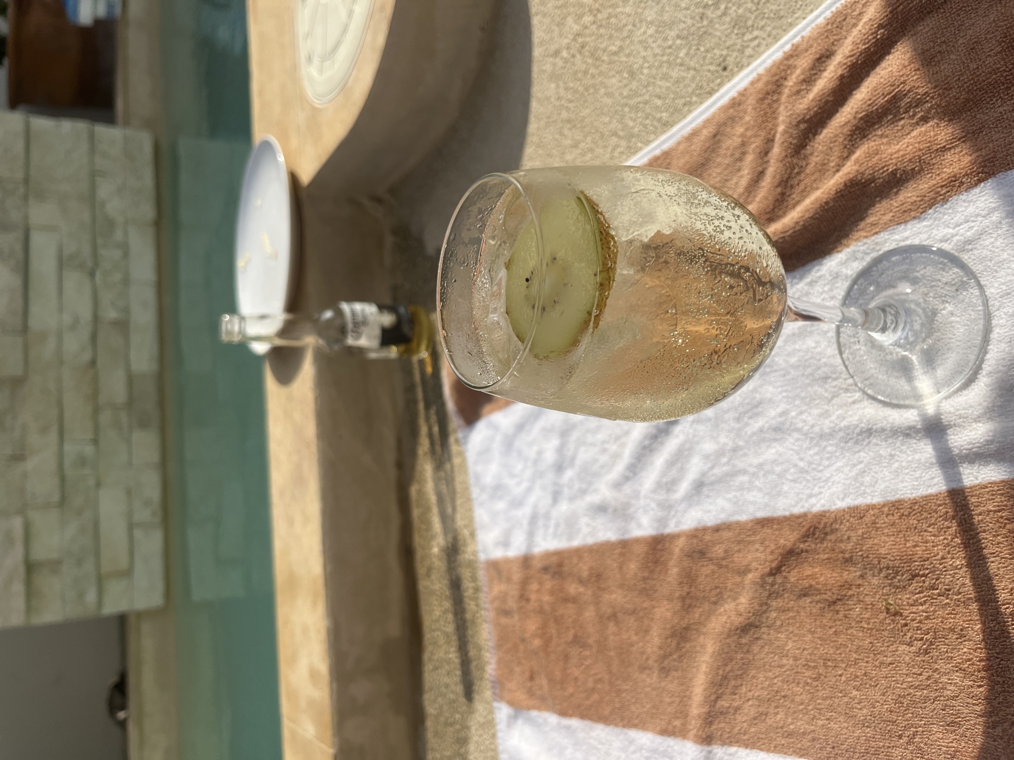Here is a description of the image:
The image shows a glass of champagne with a slice of kiwi in it, placed on a striped towel. The location appears to be outdoors, possibly by a pool. A bottle of Corona beer and a plate are also visible. The scene is set in Brisas de Zicatela, Mexico. The presence of a drink and towel suggests a relaxed, leisure-focused environment.