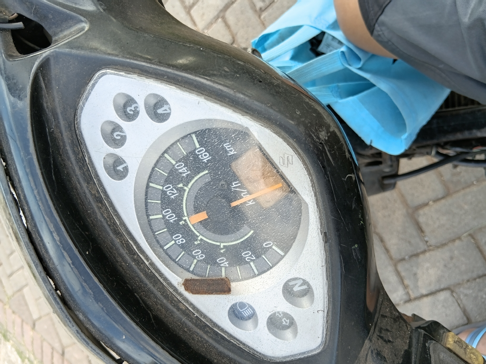 A motorcycle's speedometer is the main subject in this close-up, eye-level, outdoor shot. The speedometer dial, showing a speed of approximately 50 km/h, is centrally located within the frame. To the left of the speedometer, there are three indicator lights: one for headlights, one for turn signals, and one marked with the letter 'N' for neutral. Above and to the right of the speedometer, four circular indicators labeled 1, 2, 3, and 4 are visible. The motorcycle's black plastic housing surrounds the speedometer. A light blue reusable bag is partially visible in the lower right corner of the image, and a person's leg, clad in gray shorts, is also present. The background consists of a paved surface made of grey interlocking bricks. The scene appears to be located in Tanggul Wetan, Indonesia.