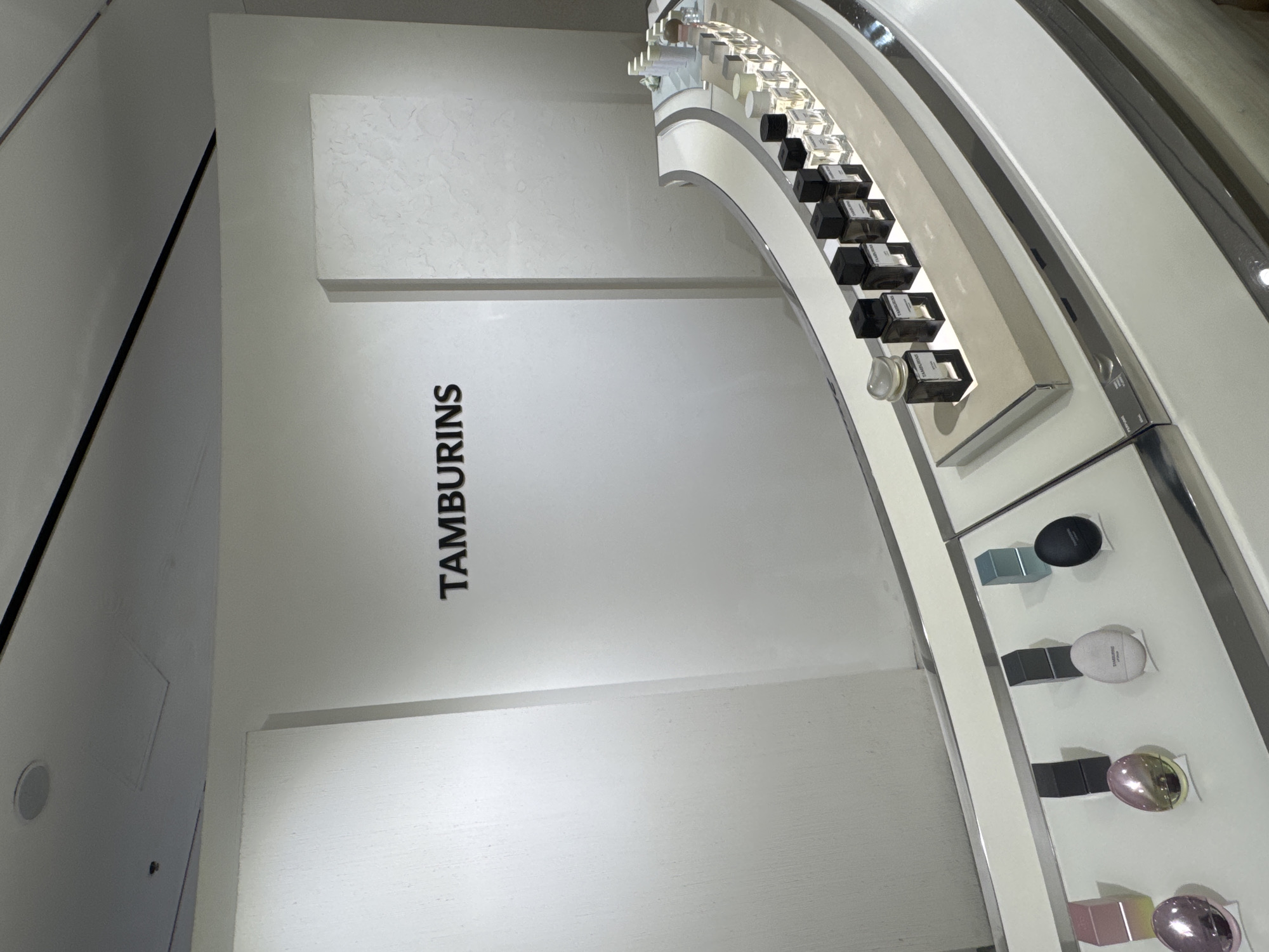 The image shows a retail display of perfume bottles and compacts, with the brand name "TAMBURINS" visible on a white wall in the background. The display is organized on a curved, illuminated shelf, with several rows of perfume bottles and smaller cosmetic items. The overall aesthetic is clean and modern, with a focus on product presentation. The scene is likely within a department store or a dedicated brand boutique. The specific location is confirmed to be within the city of Shanghai, China.