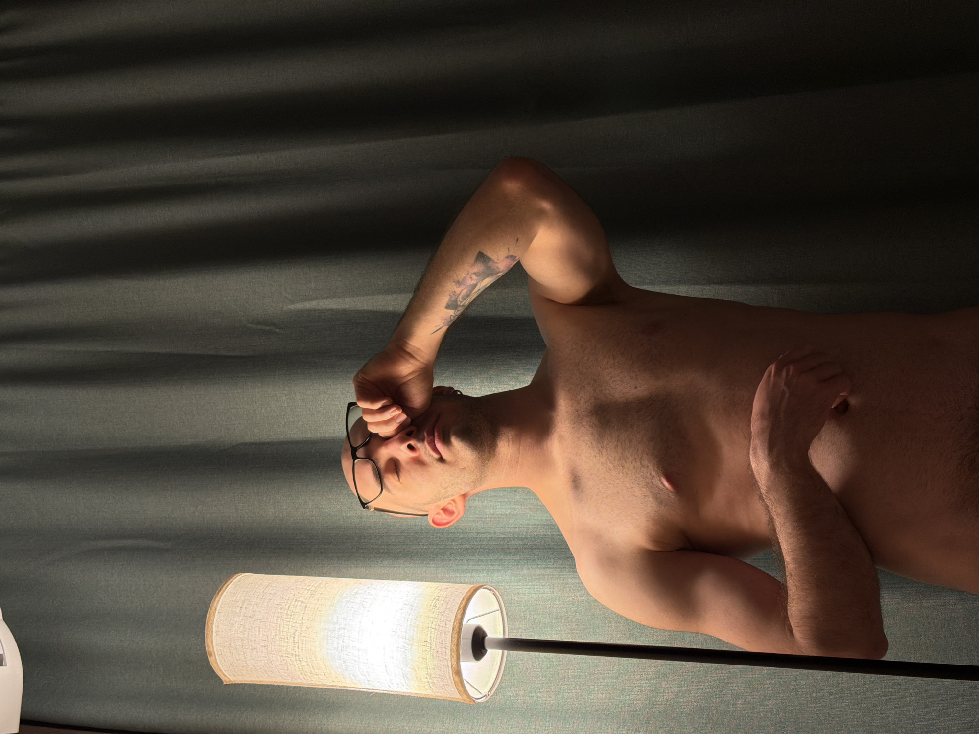 A shirtless man with glasses lies on his side in a dimly lit room. He has a tattoo on his upper left arm. A floor lamp stands beside him, casting light upwards. The scene appears to be indoors, with a wall or fabric backdrop behind the man. There is no evidence to identify the city of Kazan, Russia.