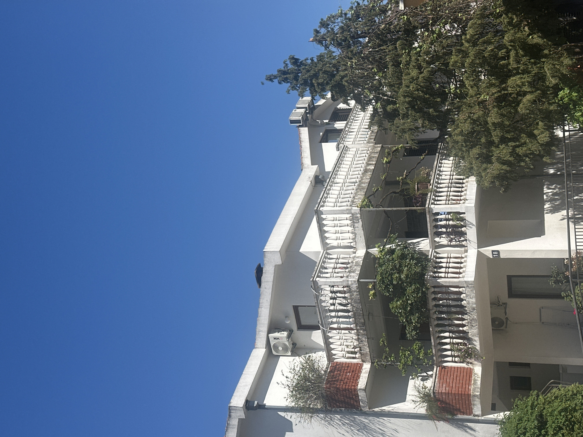 The image depicts a multi-story building in Budva, Montenegro, with a white facade and decorative balconies. The building features air conditioning units and is partially obscured by trees. The sky is clear and blue. The building's address, "18", is visible.