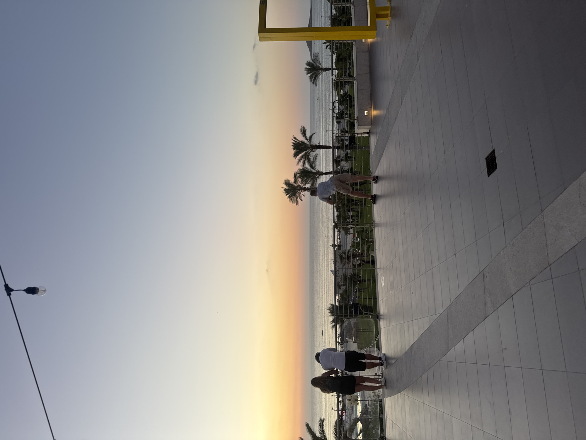 The image depicts a sunset view from a raised balcony or walkway overlooking the sea in Isola delle Femmine, Italy. In the foreground, the tiled ground has a diagonal patterned section. Three people are visible: two standing together, a woman in a black dress and a man in a white shirt and dark shorts, looking out at the sea, and a man in a light-colored shirt and tan shorts leaning against a railing. The sea stretches to the horizon, where a warm orange and yellow sunset is visible. Palm trees dot the landscape between the balcony and the sea. In the distance, a yellow structure, possibly an art installation or seating area, is present on the right side of the image. String lights hang from a wire across the top of the frame.