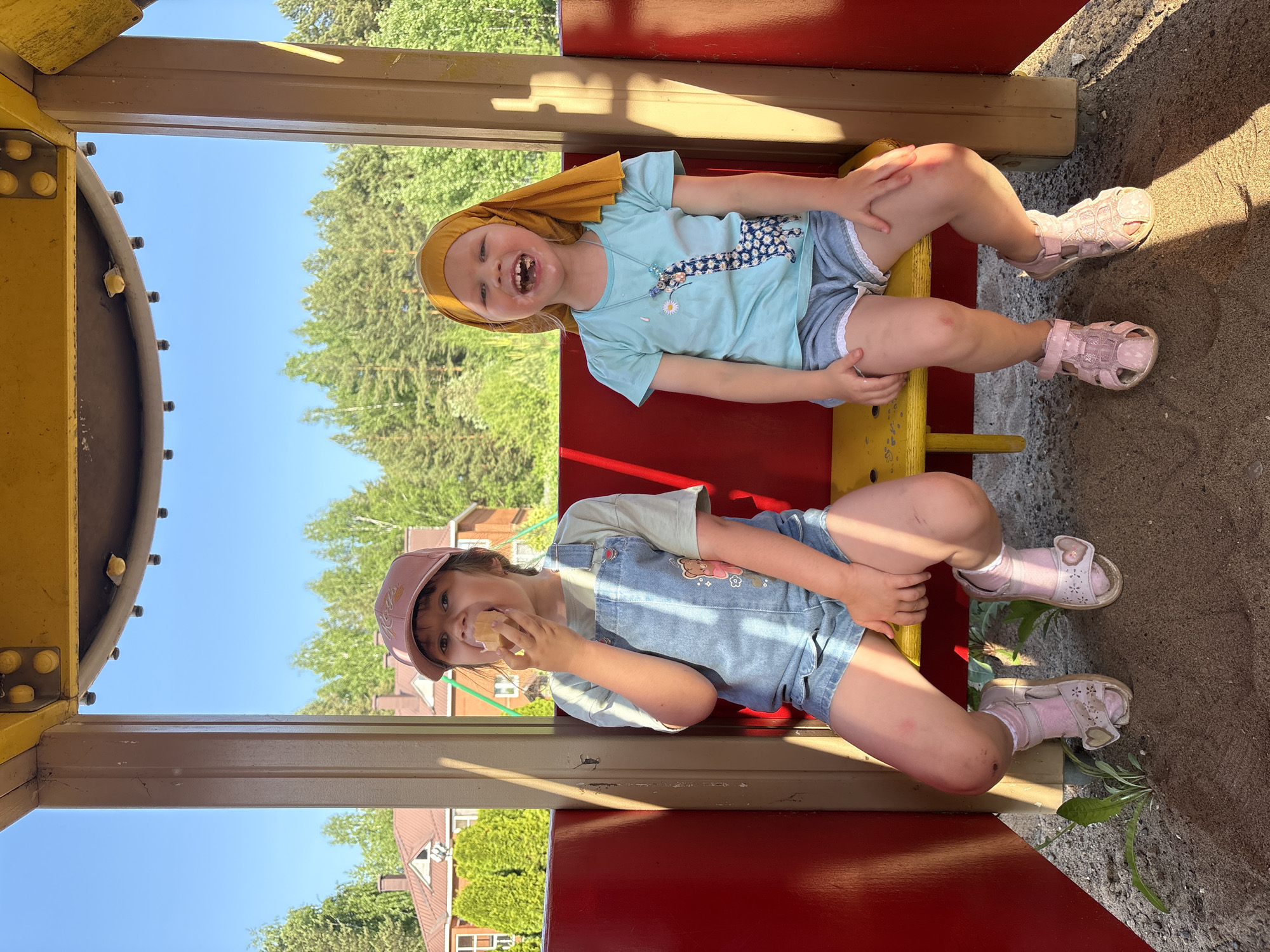 Two young girls are sitting inside a play structure, likely at a playground in Chaikovsky, Russia. The play structure has yellow and red painted components. The girl on top wears a yellow headscarf, a light blue shirt with a giraffe graphic, and light-colored shorts. She is laughing, revealing missing teeth. The girl below is wearing a pink cap, a light green t-shirt, and denim overalls. She is eating something. Both girls are wearing pink sandals. The playground appears to be surrounded by trees and grass. A building roof is visible in the background. There is sand underneath the play structure. The girls both have scrapes on their legs.