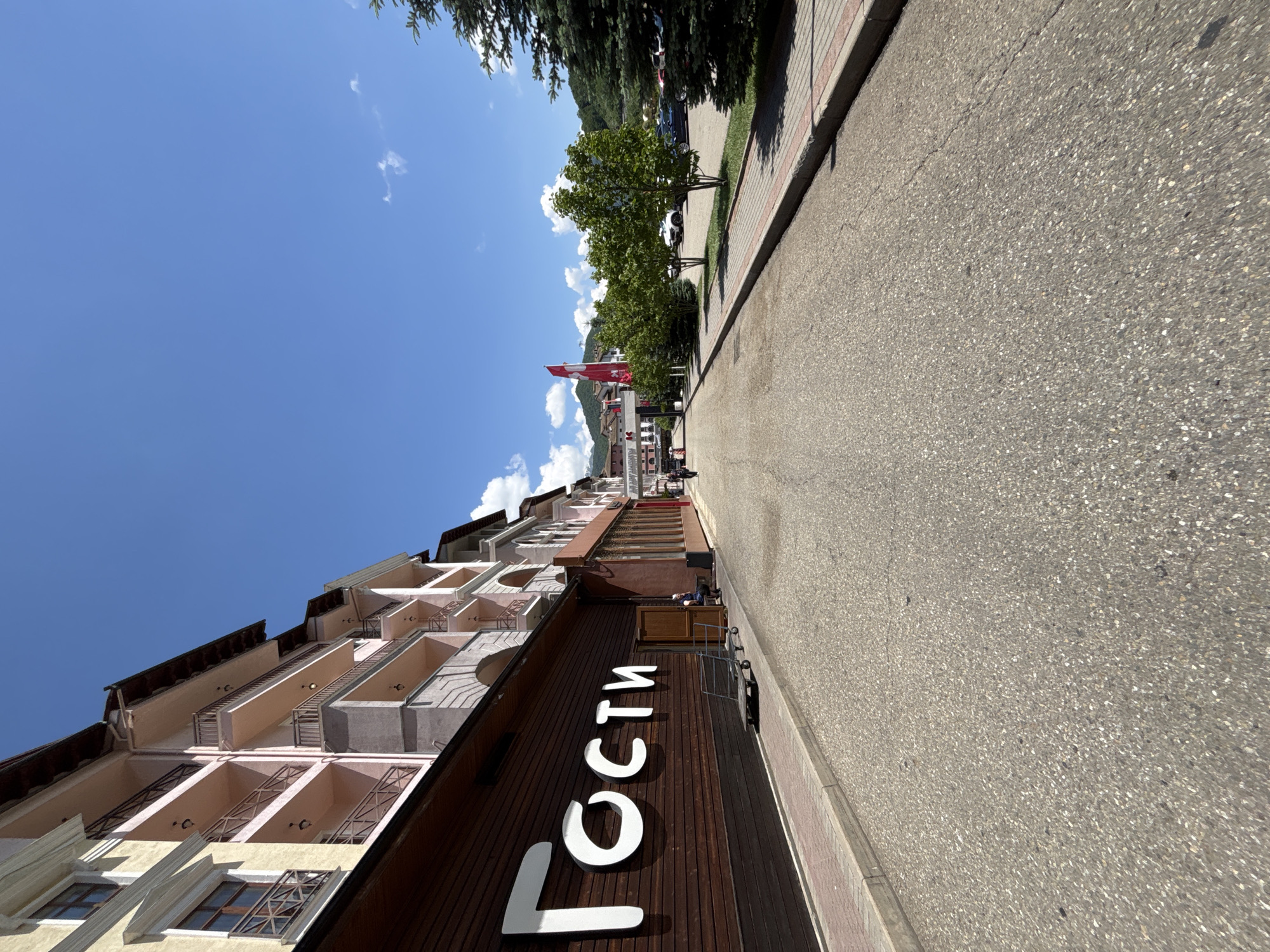 The image depicts a sunlit street scene in Estosadok, Russia, characterized by a row of multi-story buildings on the left and a paved road leading into the distance on the right. The building on the immediate left features a prominent wooden facade with large white lettering that reads "Гости" (Gosti), indicating it's likely a hotel or guesthouse. The upper floors of this building, and subsequent buildings further down the street, are painted in pastel shades of yellow and pink, with balconies and arched windows.
A person is seated on a bench outside the entrance of the building with the "Гости" sign, next to a metal shopping cart. Further down the street, another individual is walking away from the camera. On the right side of the road, a parking area is visible, with several cars parked under a bright blue sky dotted with a few white clouds. Trees, including evergreen and deciduous varieties, are planted along the sidewalk and in the parking area. In the background, rolling hills covered in green foliage rise under the clear sky, suggesting a mountainous or hilly terrain surrounding the town. A large red flag with a white cross, possibly representing Switzerland, is prominently displayed on a flagpole in the middle ground. The overall atmosphere is one of a serene, sunny day in a resort town.