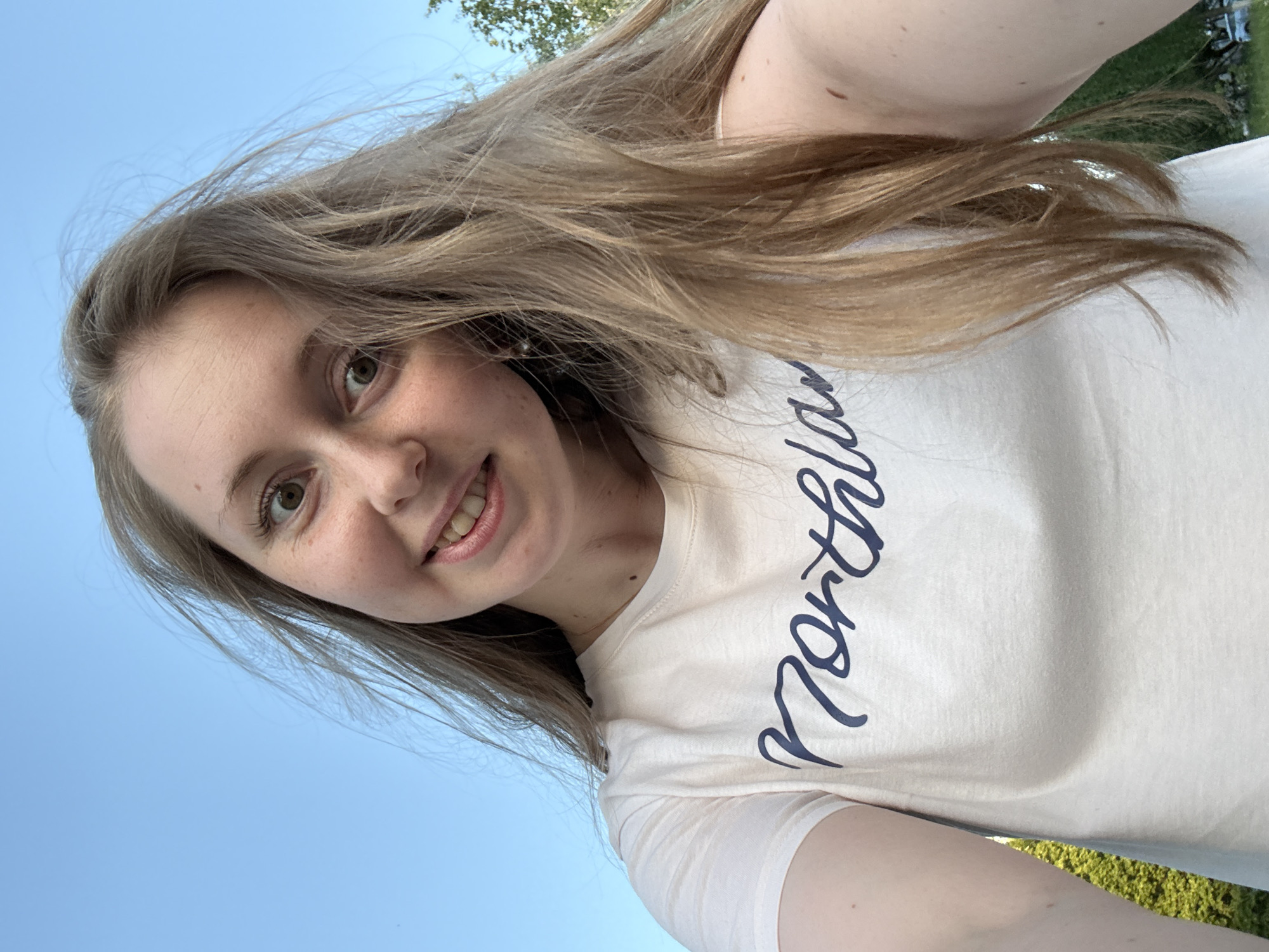 A young woman with light brown hair, fair skin, and light brown eyes is taking a selfie outdoors. She is wearing a white t-shirt with the word "Northland" in blue script. The sky is visible in the background, appearing blue. Some trees are visible in the upper right corner and the bottom of the frame, suggesting an outdoor setting. The image is tilted.