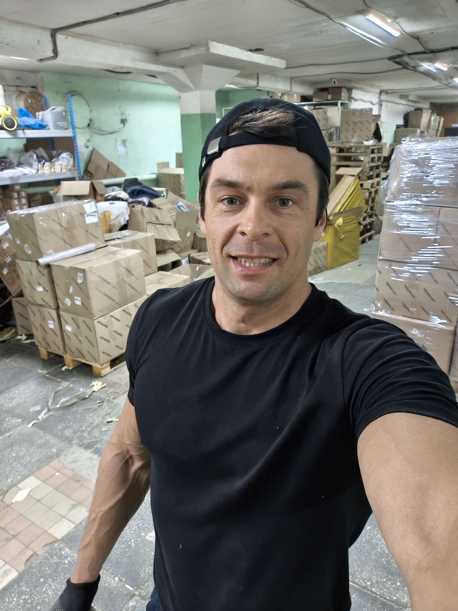 A man in a black t-shirt and black cap stands in what appears to be a warehouse or storage facility. He is taking a selfie. The background consists of pallets stacked with cardboard boxes. Some boxes are wrapped in clear plastic. The ceiling is industrial with exposed pipes and lights. The floor is tiled in a geometric pattern. Additional cardboard boxes and shelves with various items are visible in the background. There is no immediate indication of the city or country.