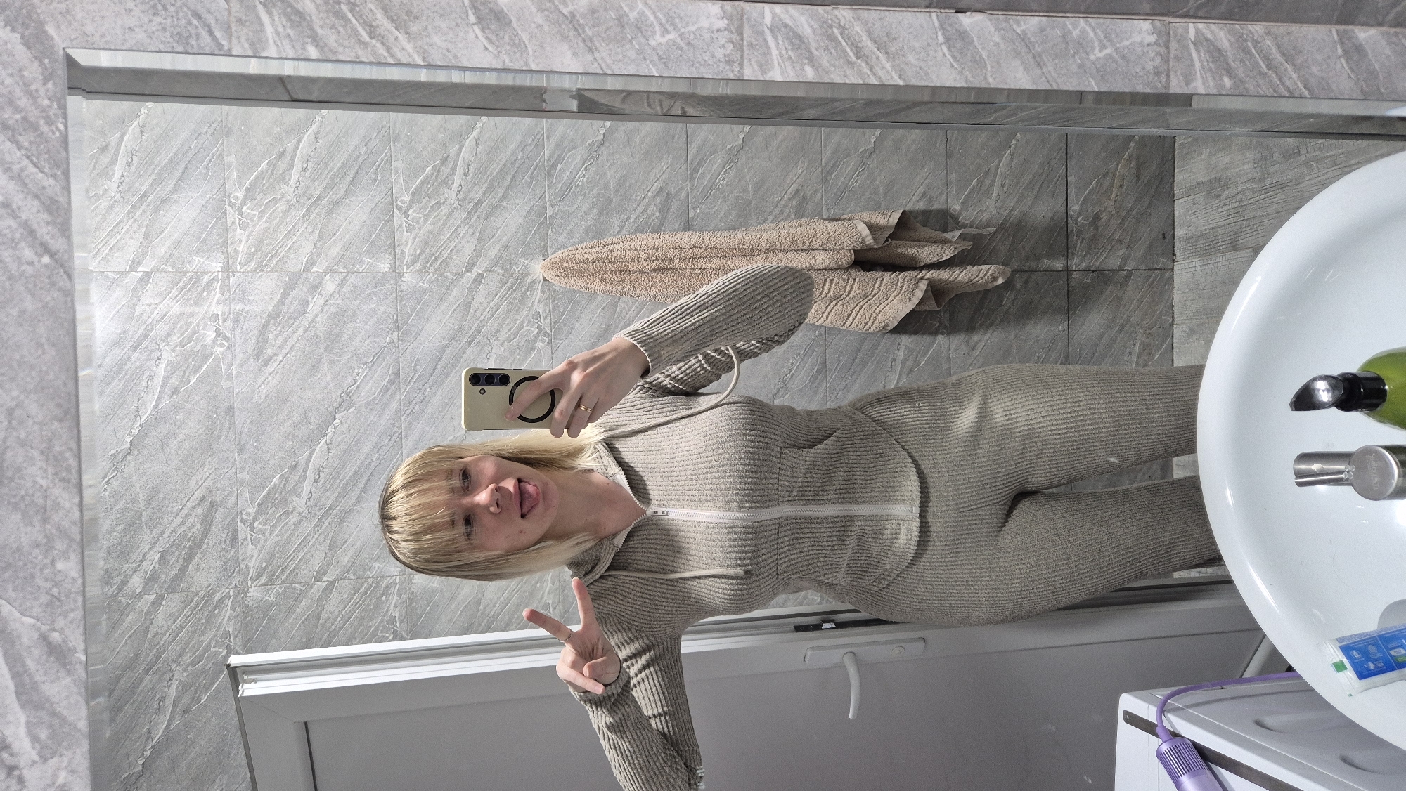 A young woman is taking a selfie in a bathroom. She is standing in front of a mirror, holding a smartphone in her right hand. She has blonde hair, is wearing a grey ribbed jumpsuit and sticking out her tongue while making a peace sign with her left hand. The bathroom has grey tiled walls, a towel hanging on the wall, a white sink, and a washing machine visible below. The photo doesn't provide enough information to identify the precise location within Batumi, Georgia.