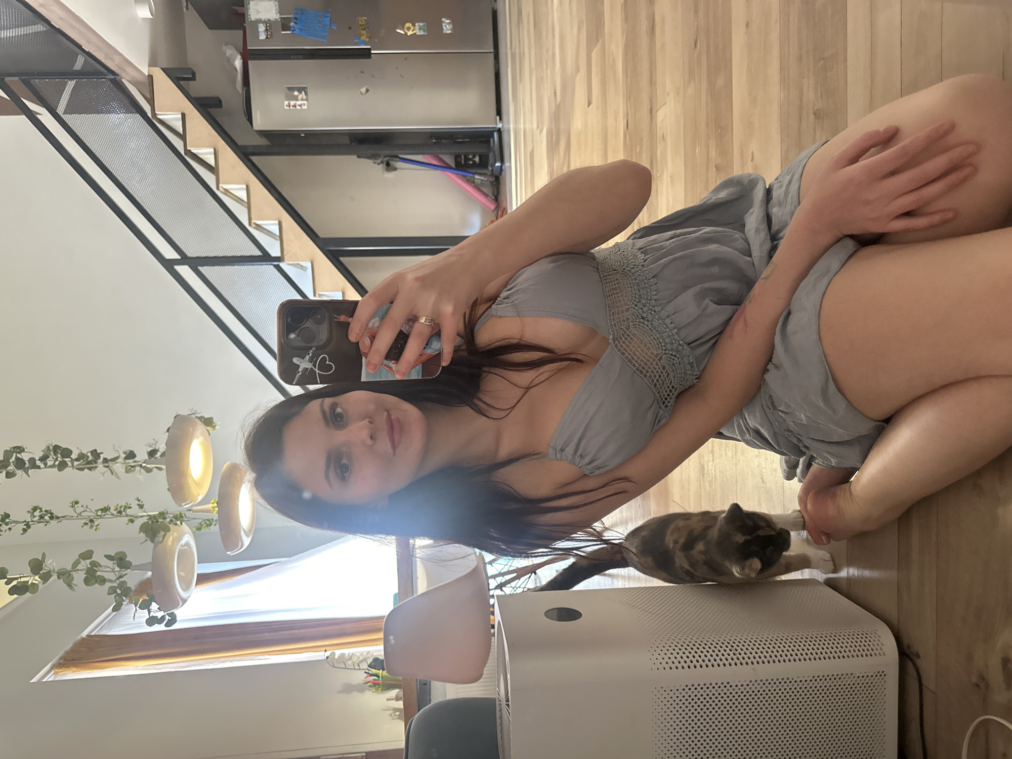 A young woman with dark hair is taking a selfie in a room. She is wearing a light blue dress and sitting on the floor. Next to her is a small calico cat. The room appears to be a loft-style apartment, with a staircase visible in the background and a refrigerator. The woman is holding a phone in her right hand, and a ring is visible on her left hand. The scene is illuminated by natural light and overhead lighting fixtures.