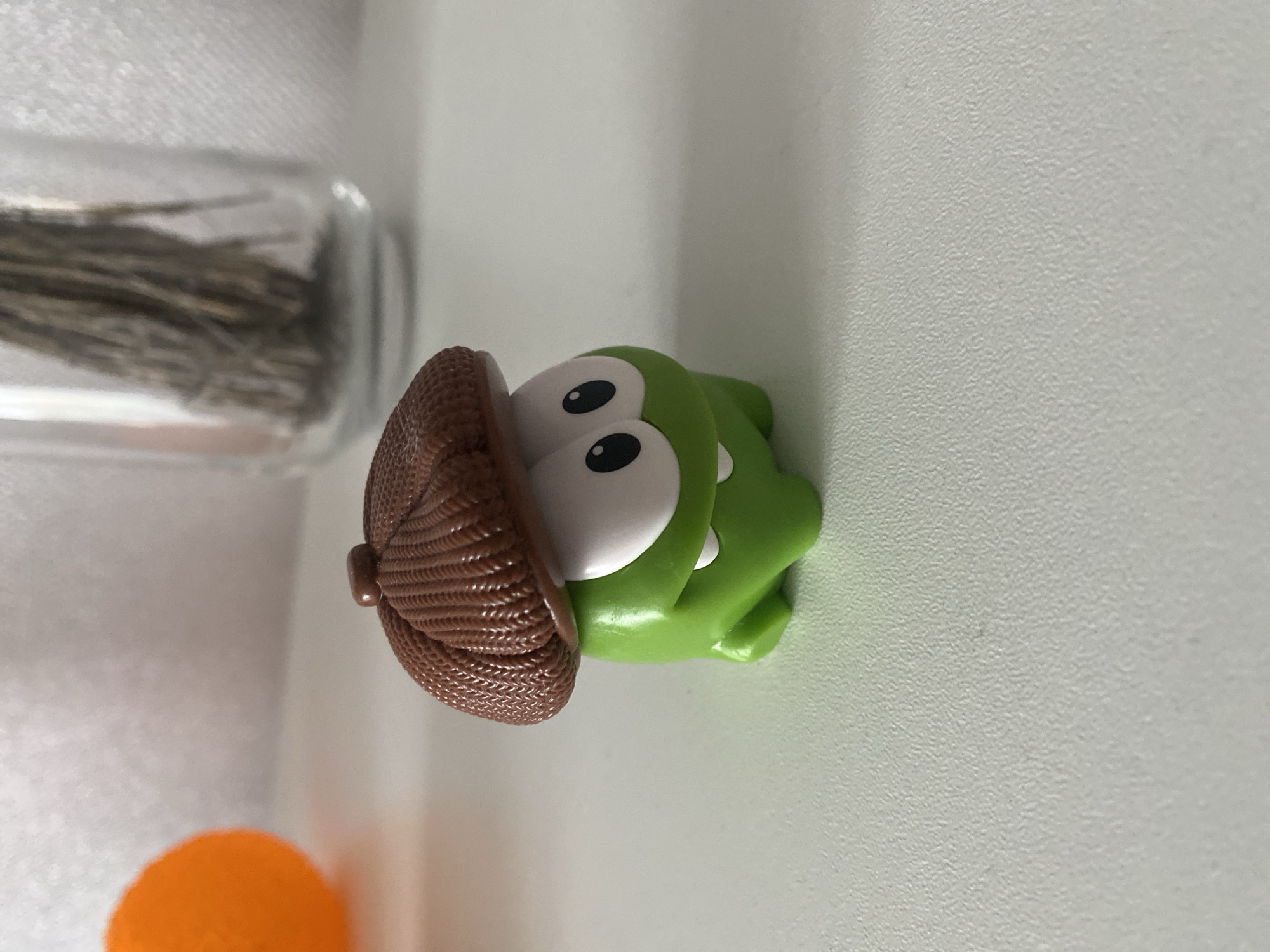 A small, green plastic toy figure, resembling Om Nom from the "Cut the Rope" franchise, sits prominently on a light gray, textured surface. The figure features large white eyes with black pupils and two small white teeth. It wears a brown, textured, dome-shaped hat with a small knob on top, positioned partially over its eyes. In the blurred background to the upper left, a vibrant orange spherical object is visible. To the upper right, a clear glass vessel containing multiple slender, dry, brown botanical elements stands in the background. The lighting is bright and even, casting subtle shadows beneath the toy. The scene is static, captured in an indoor environment, at the location of Unknown, Unknown.