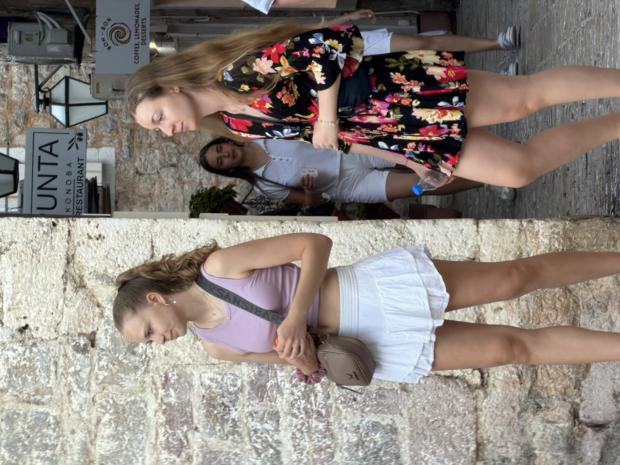 In Budva, Montenegro, two young women stand on a cobblestone street with rough-hewn stone walls. The woman on the left, with her hair in a ponytail, wears a lavender tank top and a white ruffled skirt, carrying a small brown crossbody bag. She holds an orange object in her hands and looks to her left. To her right, another young woman with long blonde hair wears a black floral dress and a black fanny pack, holding a clear plastic water bottle. Behind them, a third young woman in a white polo shirt and white shorts, and another person are partially visible. Signs for "UNTA KONDBA RESTAURANT" and "BON-BON COFFEE, LEMONADES, DESSERTS" are mounted on the stone wall. A street lamp is also visible.